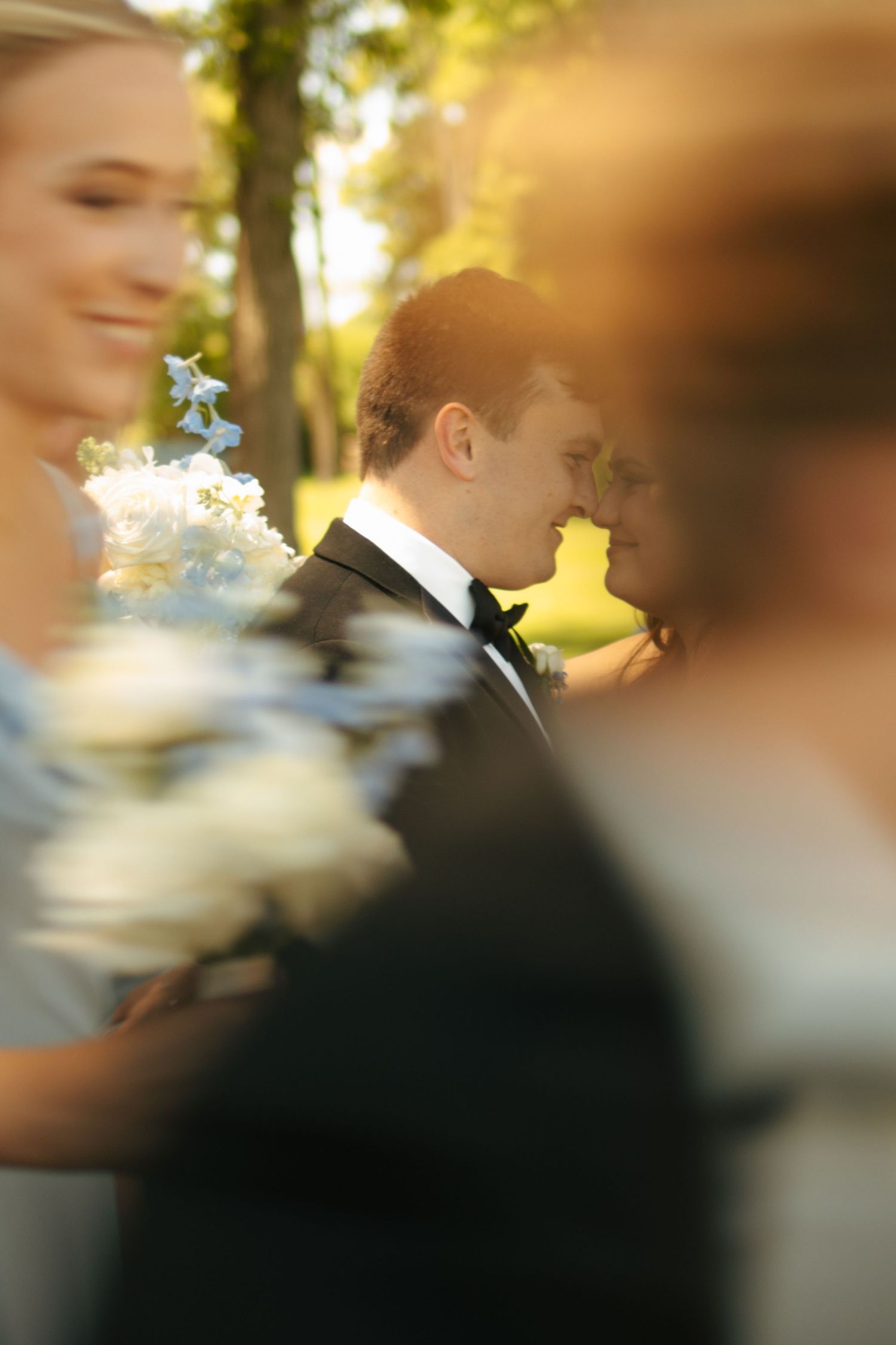 Groom in black tuxedo stands outdoors in golden sunlight, surrounded by wedding party members holding white floral bouquets.