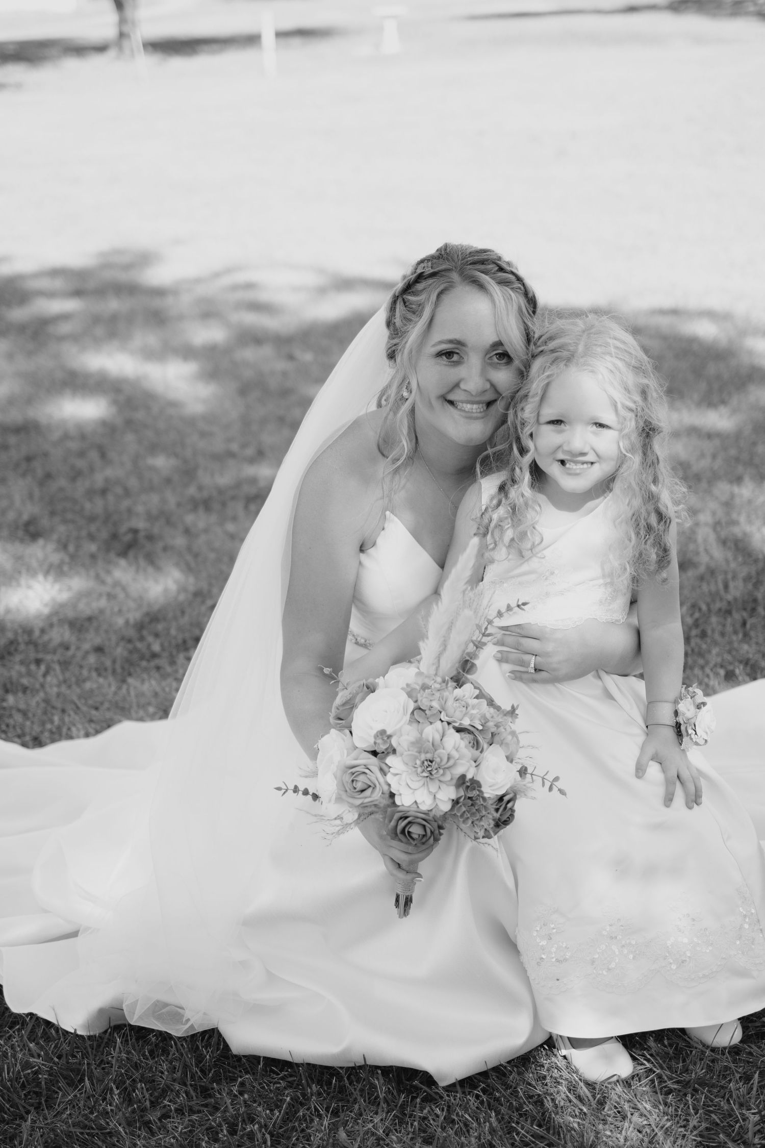 Bride in white gown and veil sits with smiling flower girl in white dress, holding bouquet, black and white wedding photo.