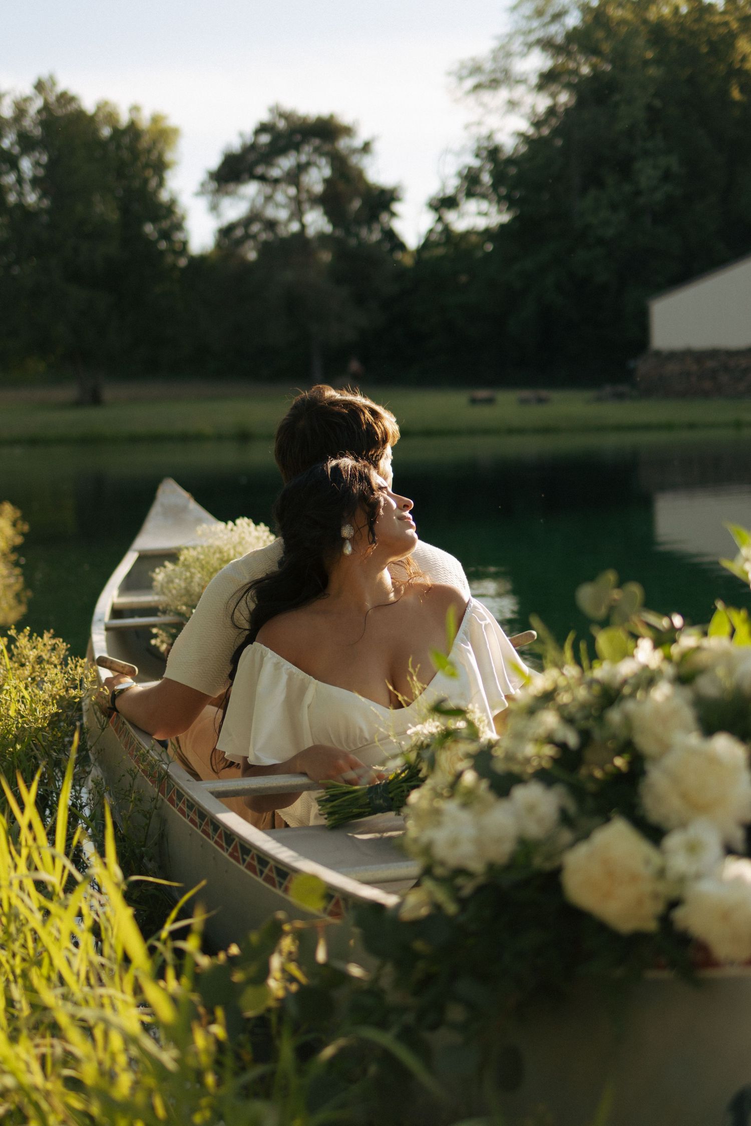 A bride and groom embrace in a floral-decorated canoe on a calm lake at golden hour.