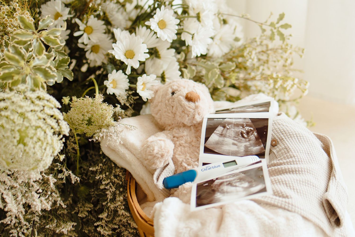 Teddy bear with ultrasound photos, pregnancy test, and white flowers arranged on a cozy knit blanket.