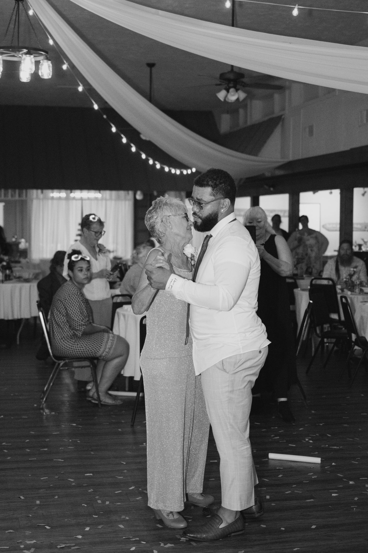 Groom shares emotional dance with elderly woman in elegant barn reception hall with string lights.