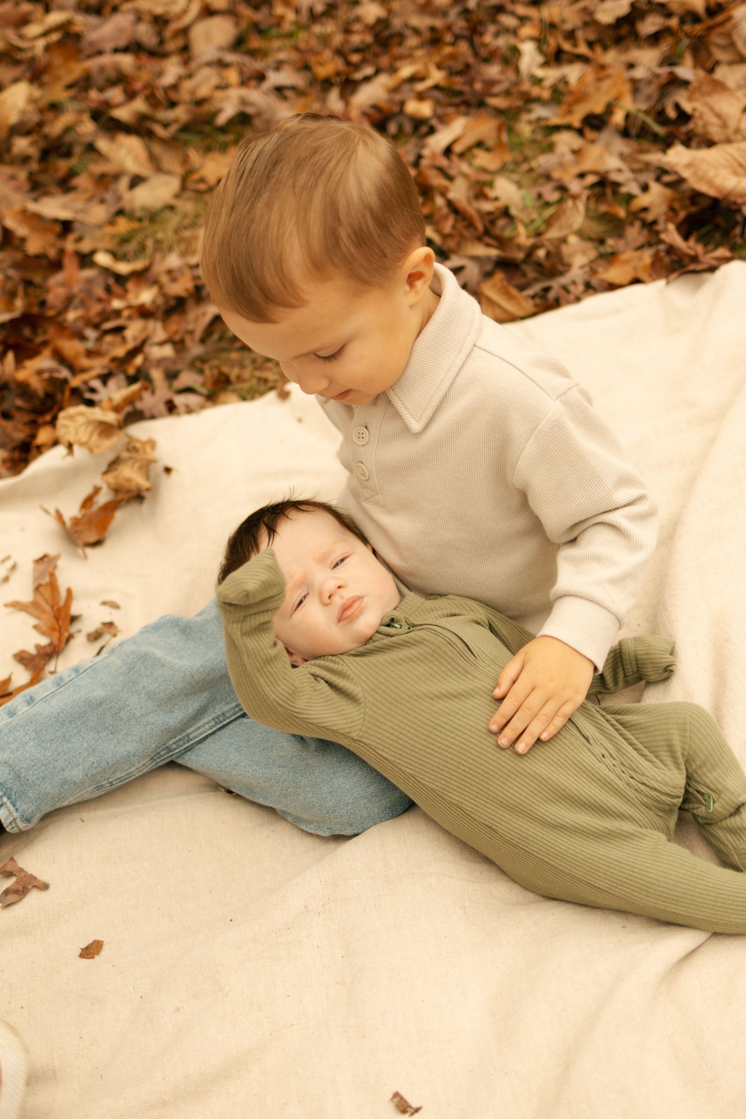 Toddler boy in beige sweater sitting with sleeping newborn baby in green outfit on white blanket surrounded by autumn leaves.