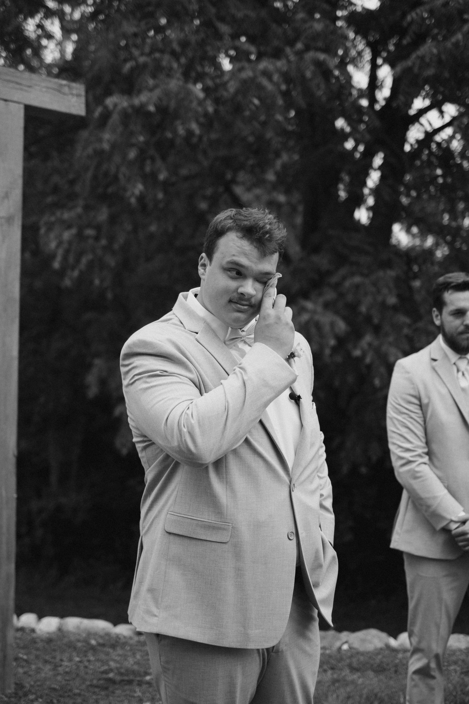 Emotional groom in light gray suit wipes tear from eye during outdoor wedding ceremony in black and white photo.