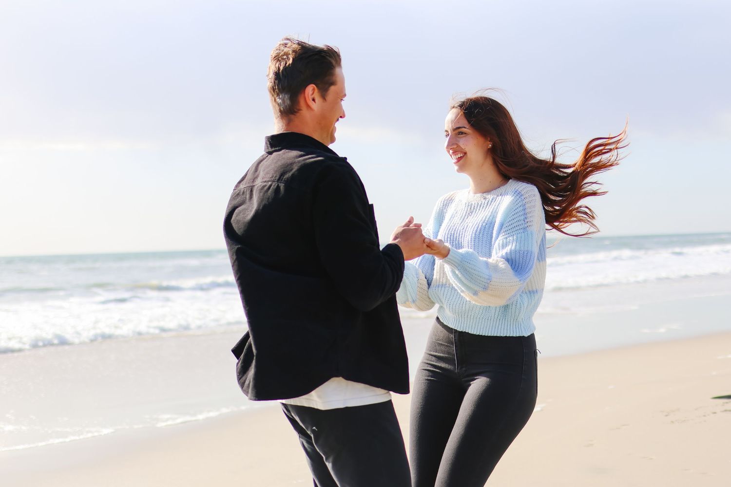 Couple holding hands and smiling on a beach with ocean waves in the background on a cloudy day.