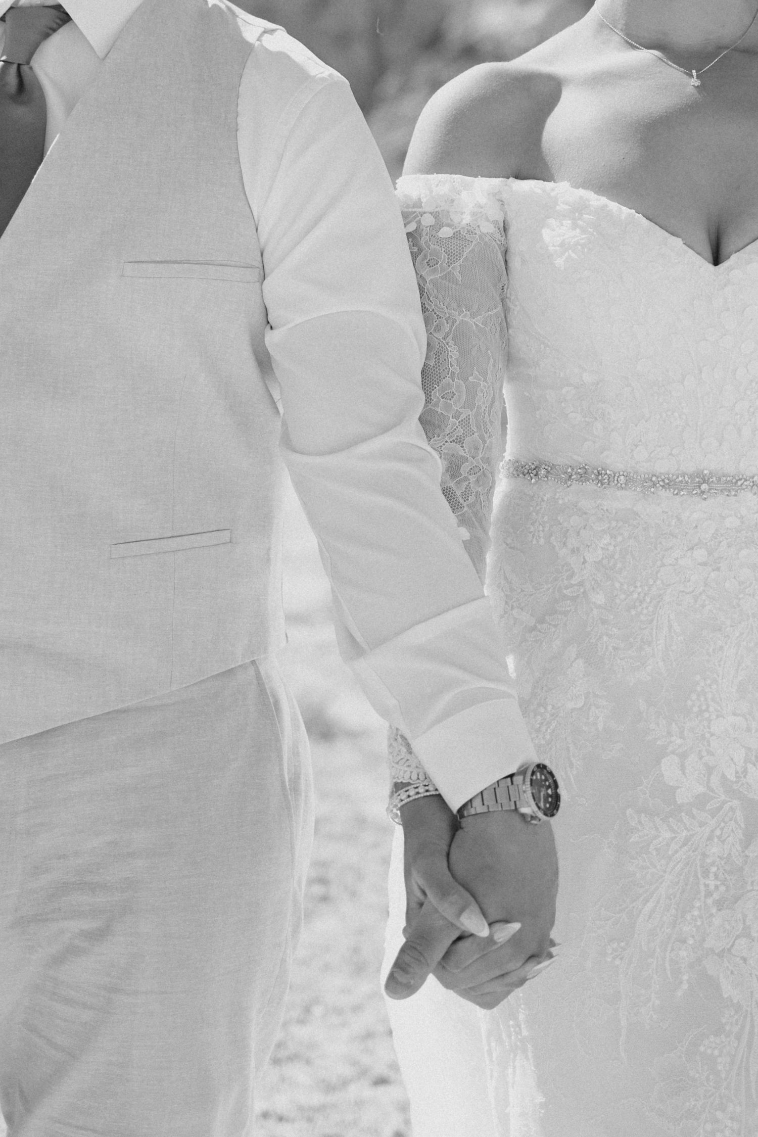 Black and white photo showing a couple holding hands, with the groom in a white vest and bride in a lace dress.