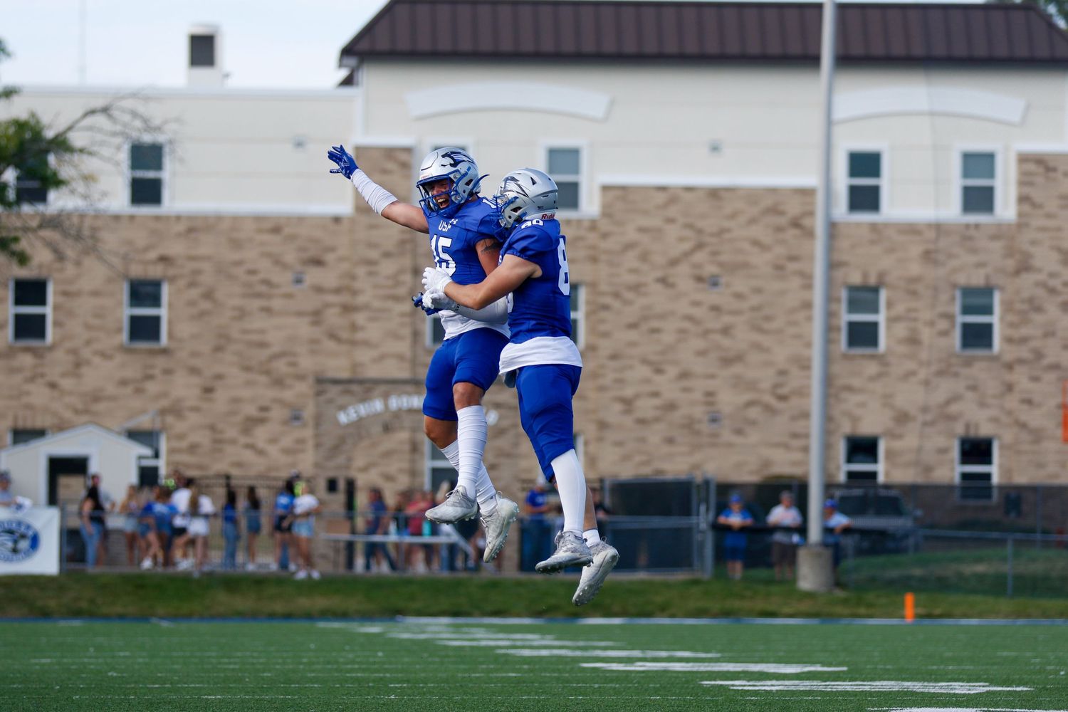 Two football players in blue uniforms leap and celebrate after making a play on the field.