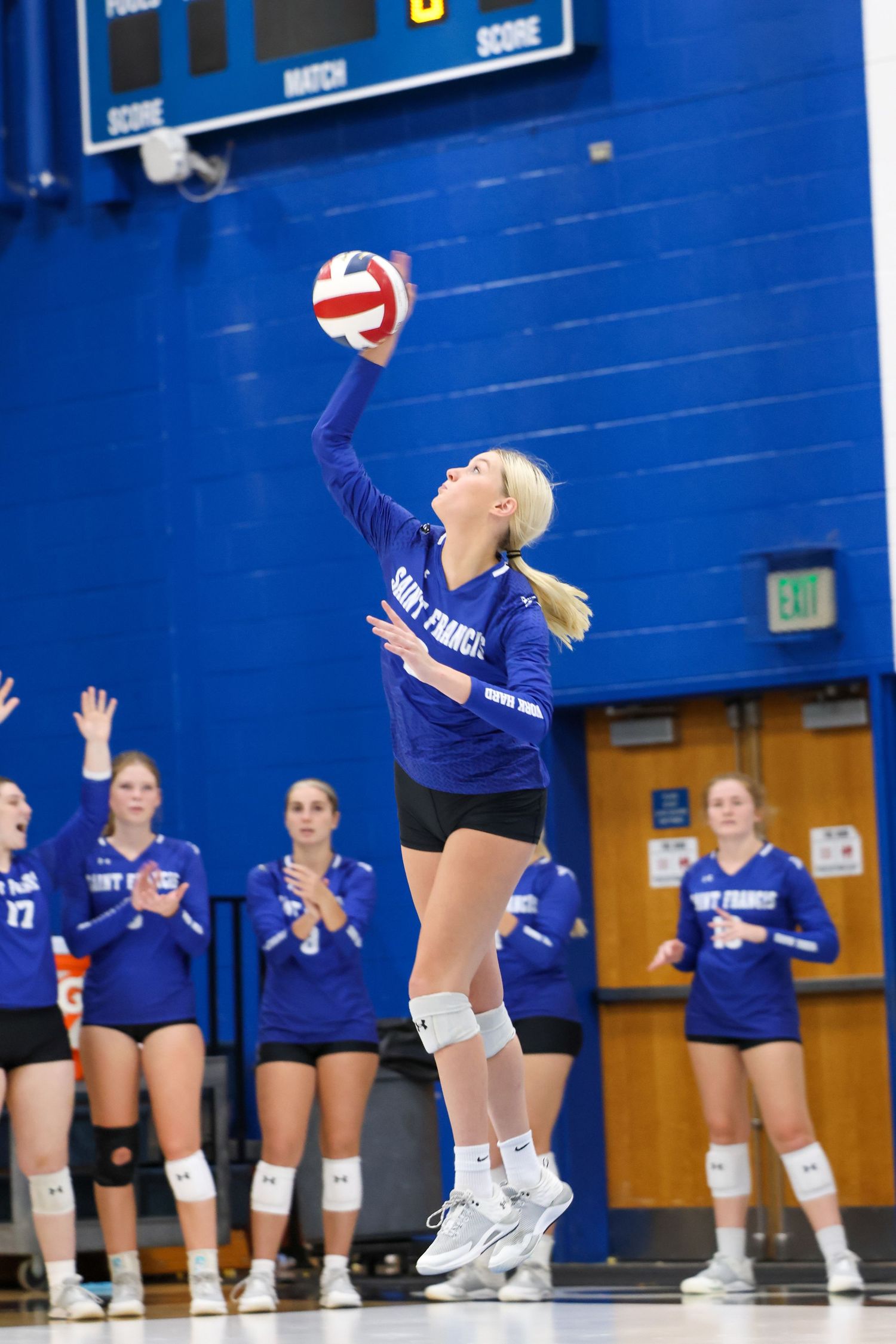 High school volleyball player in navy blue uniform jumps to serve ball during match in gym with blue walls.