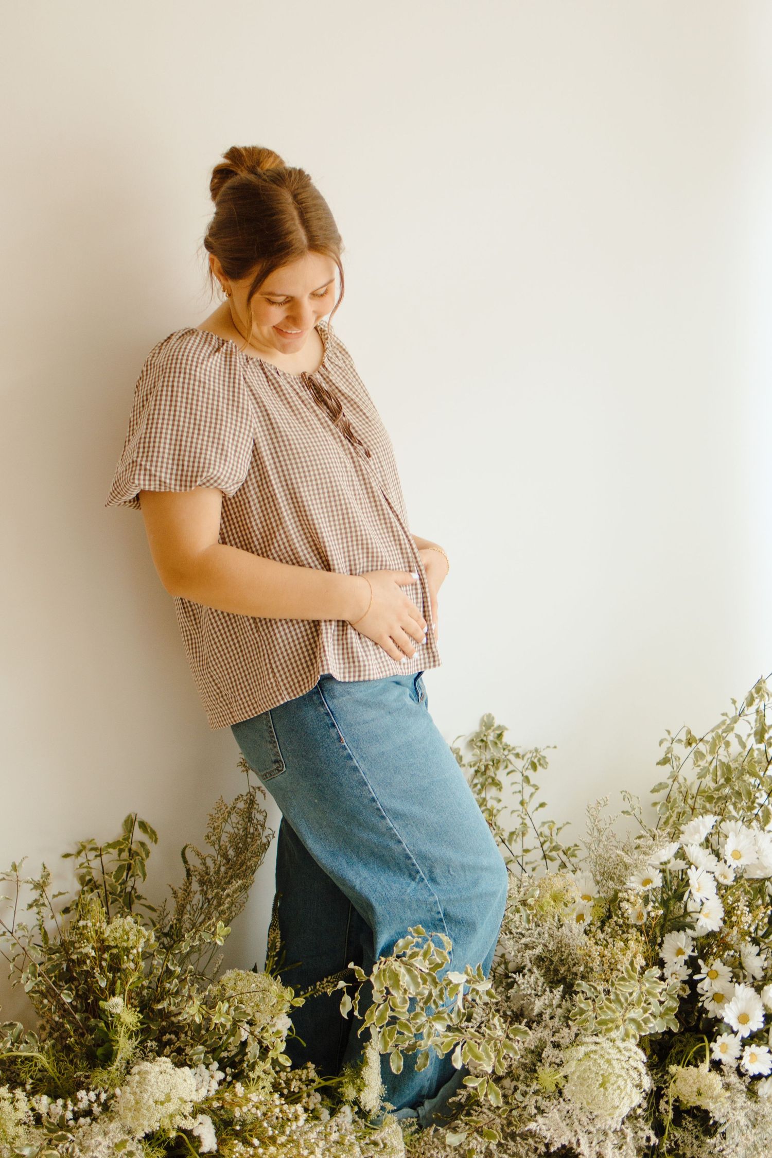Pregnant woman in plaid top and jeans smiling down at baby bump, surrounded by white and green flowers.