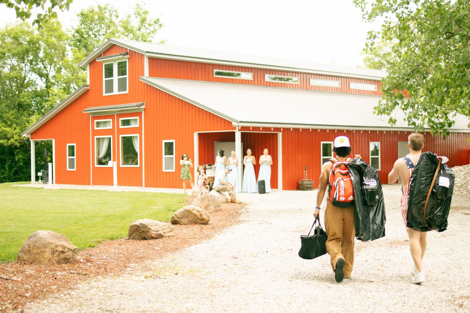 Two men carry bags toward a red barn wedding venue on a gravel path surrounded by lush green trees.