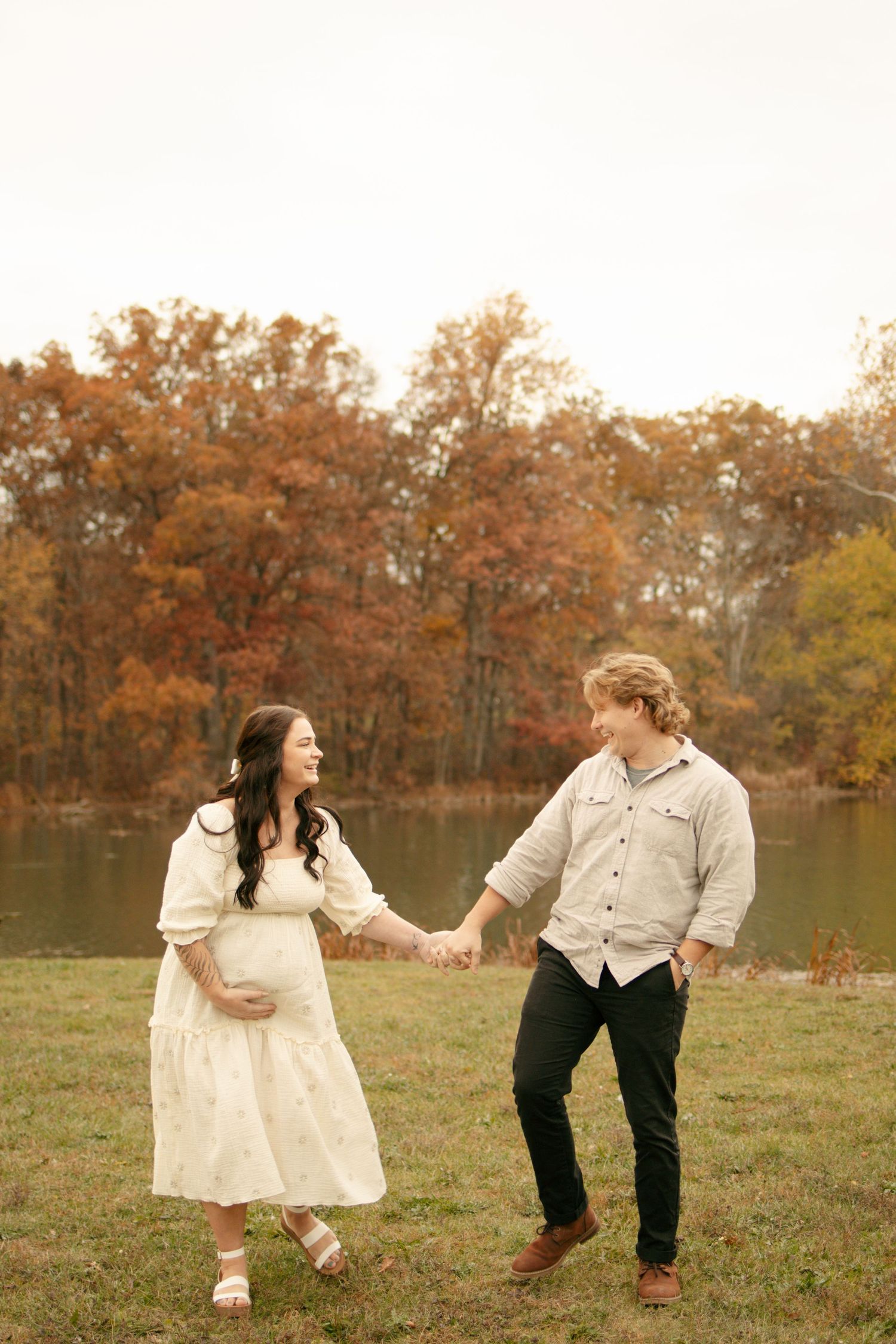 Pregnant couple holding hands by a serene autumn lake, woman in white maternity dress, colorful fall trees behind them.