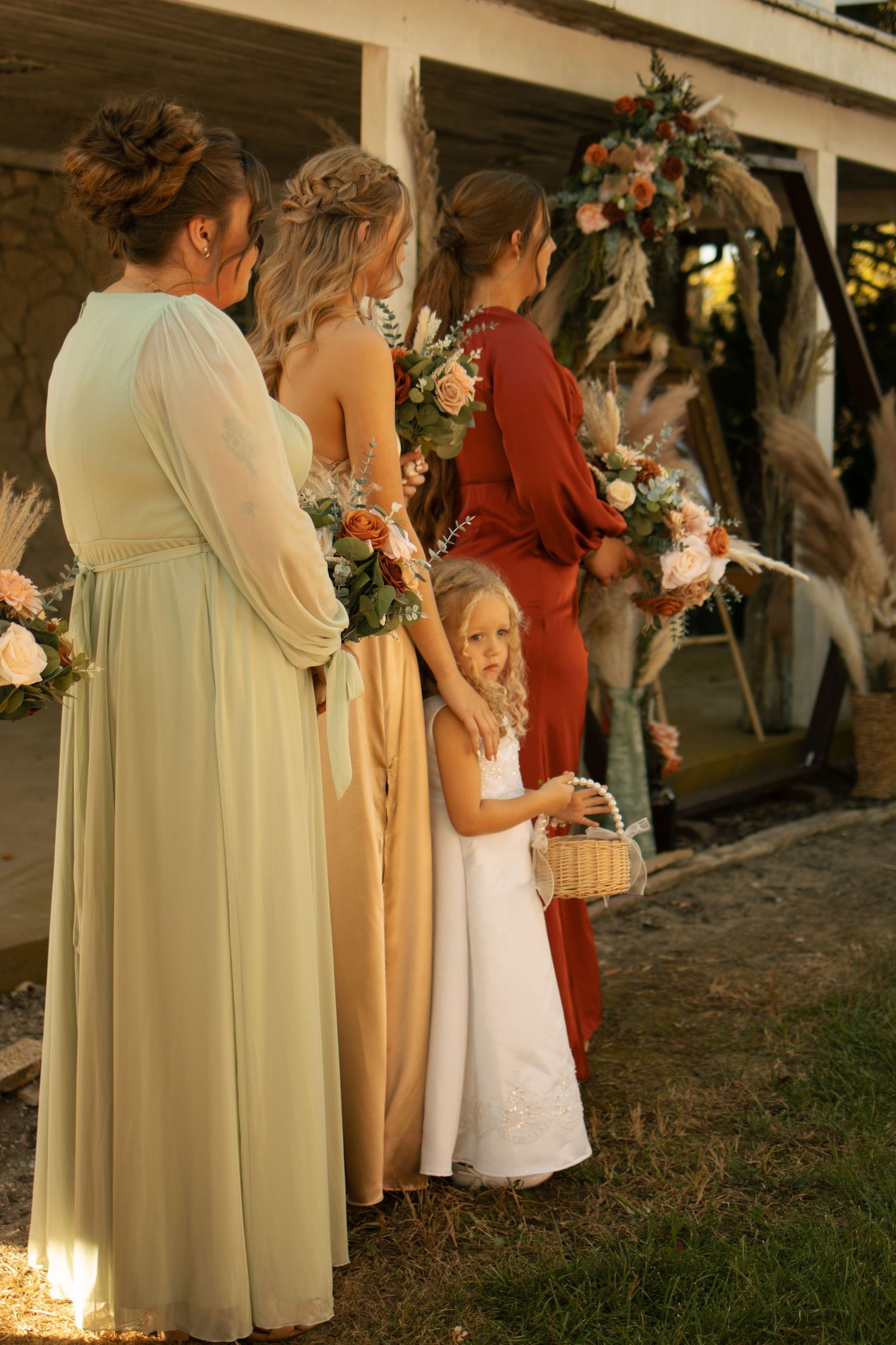 Bridesmaids in colorful gowns hold bouquets beside a flower girl in white at an outdoor wedding ceremony.