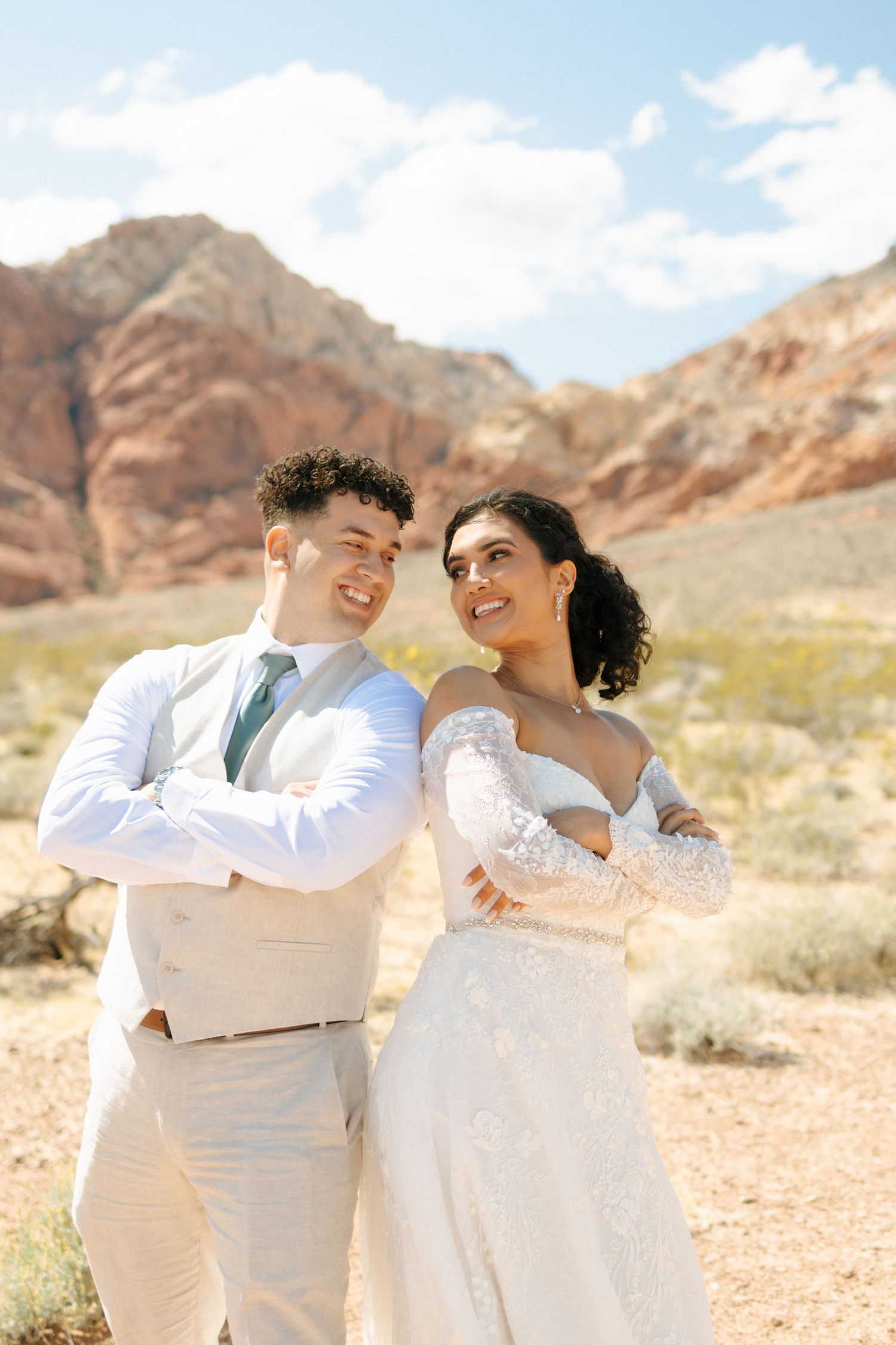 A couple in white attire share joyful moments against a desert mountain backdrop with red rocks.