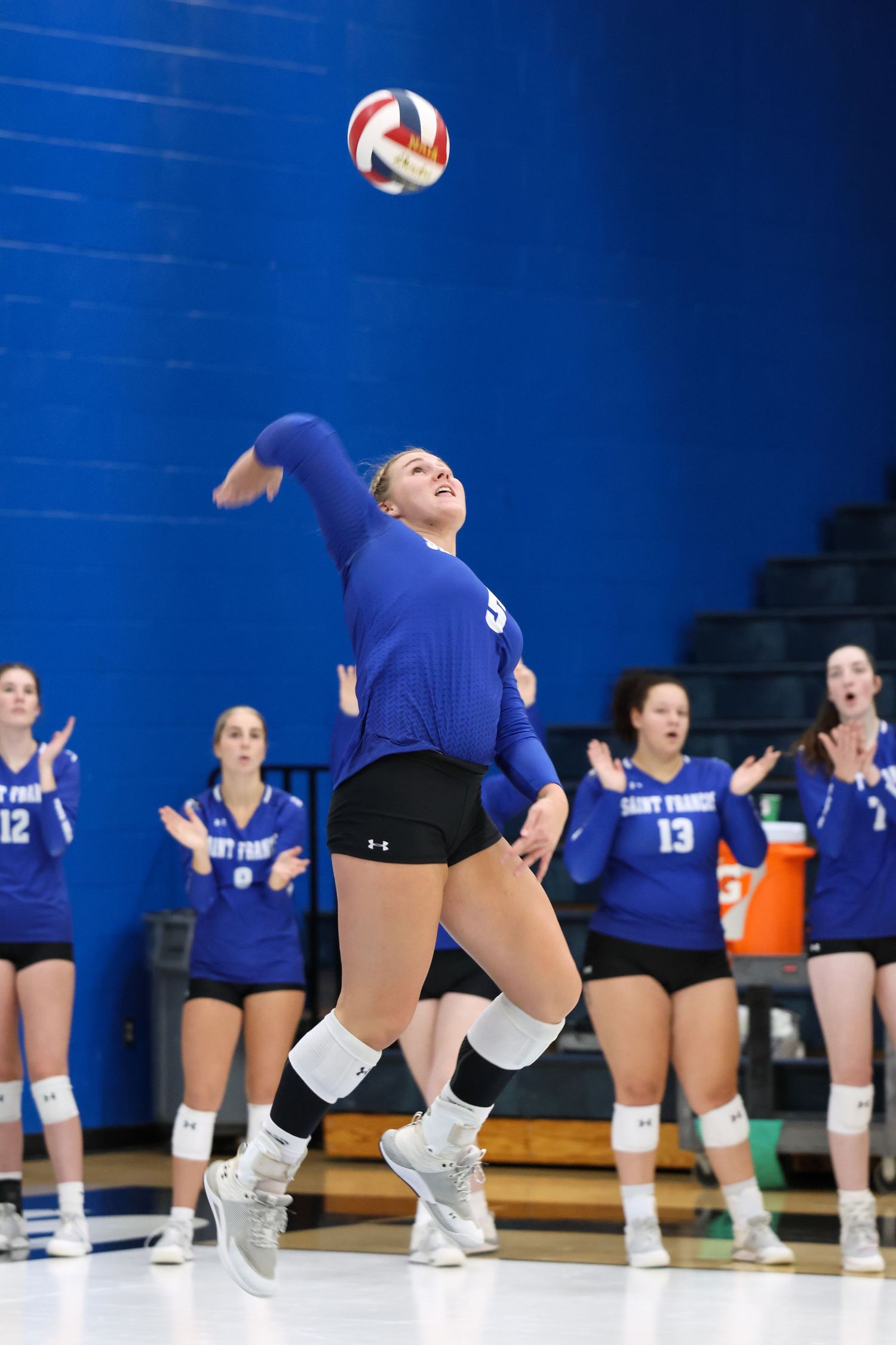Volleyball player in blue uniform jumps to hit ball during indoor match with teammates visible in background.