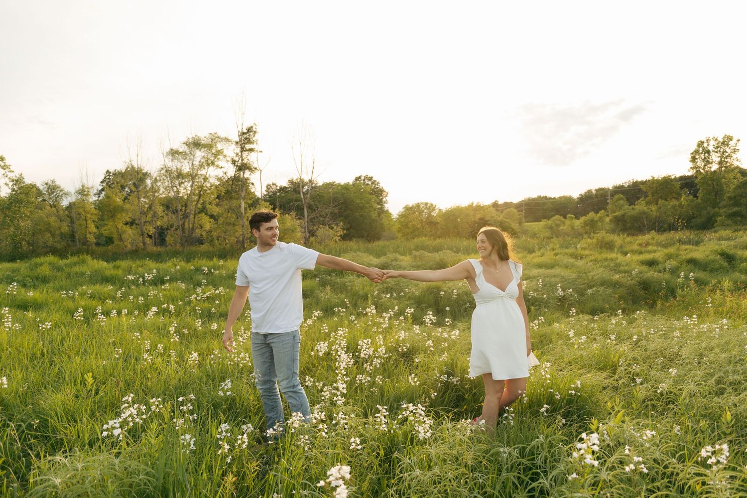 Couple holding hands in a sunlit wildflower meadow, woman in white dress, man in white shirt and jeans, golden hour.