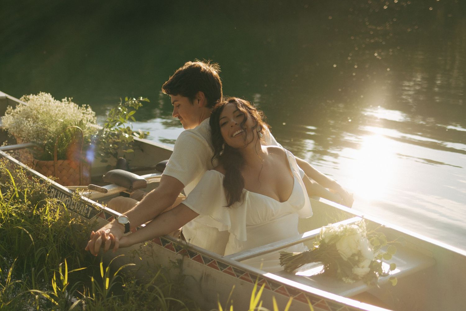 A couple in wedding attire shares a romantic moment in a sunlit rowboat surrounded by lush greenery and water.