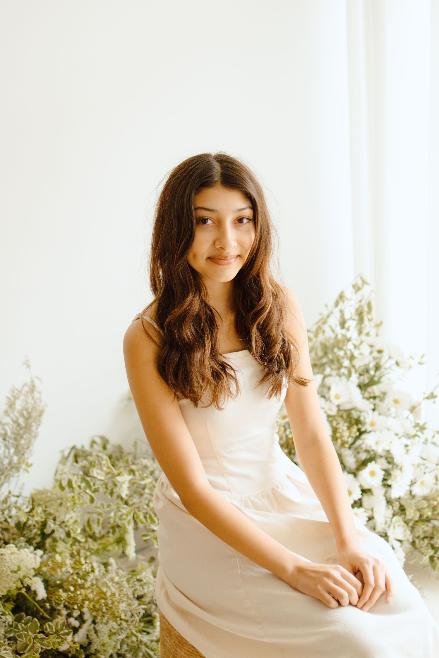 Young woman in white dress sitting among white flowers and greenery in a bright, airy studio setting.