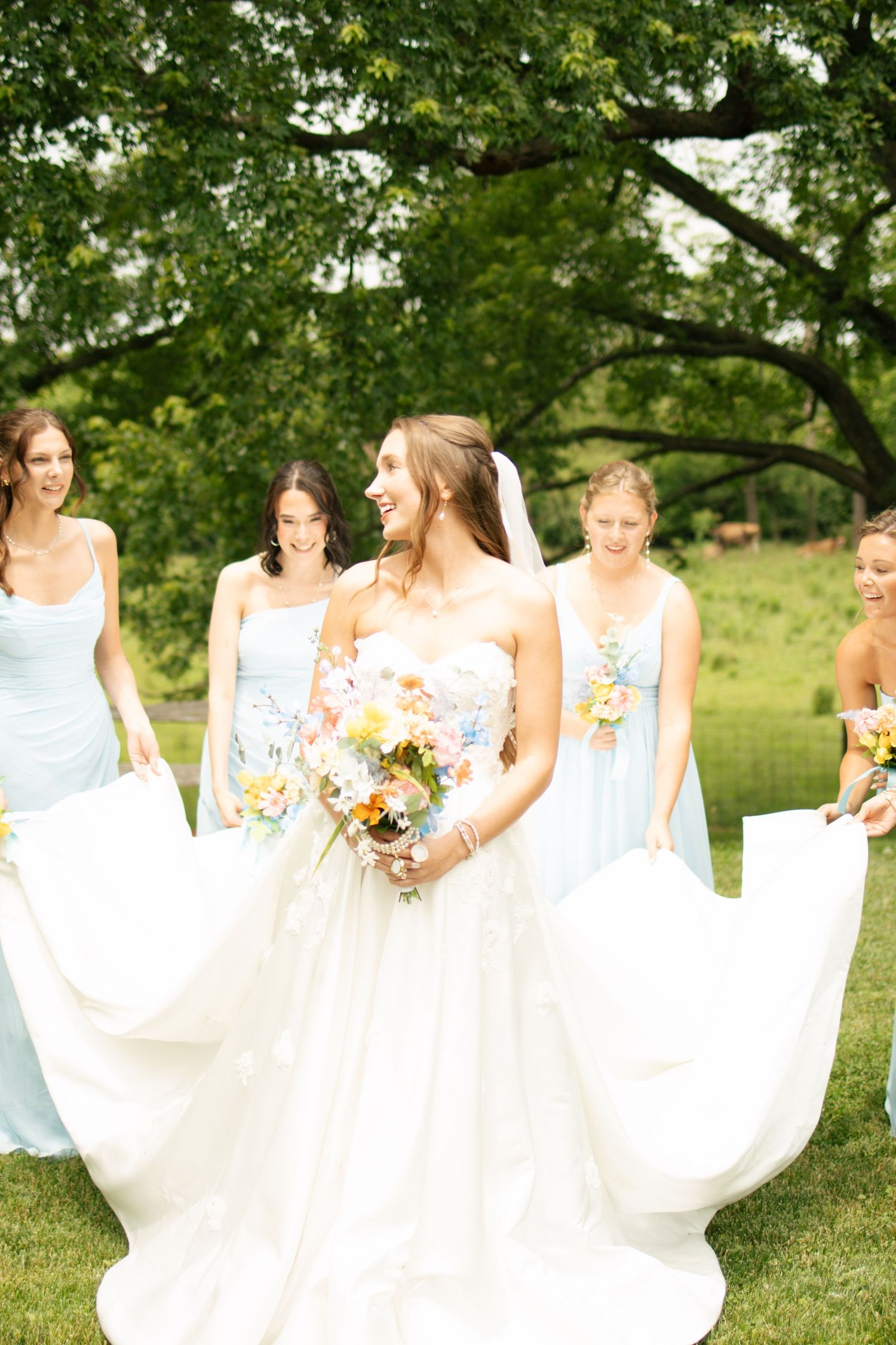 Bride in white gown walks with bridesmaids in light blue dresses outdoors, holding colorful bouquet.
