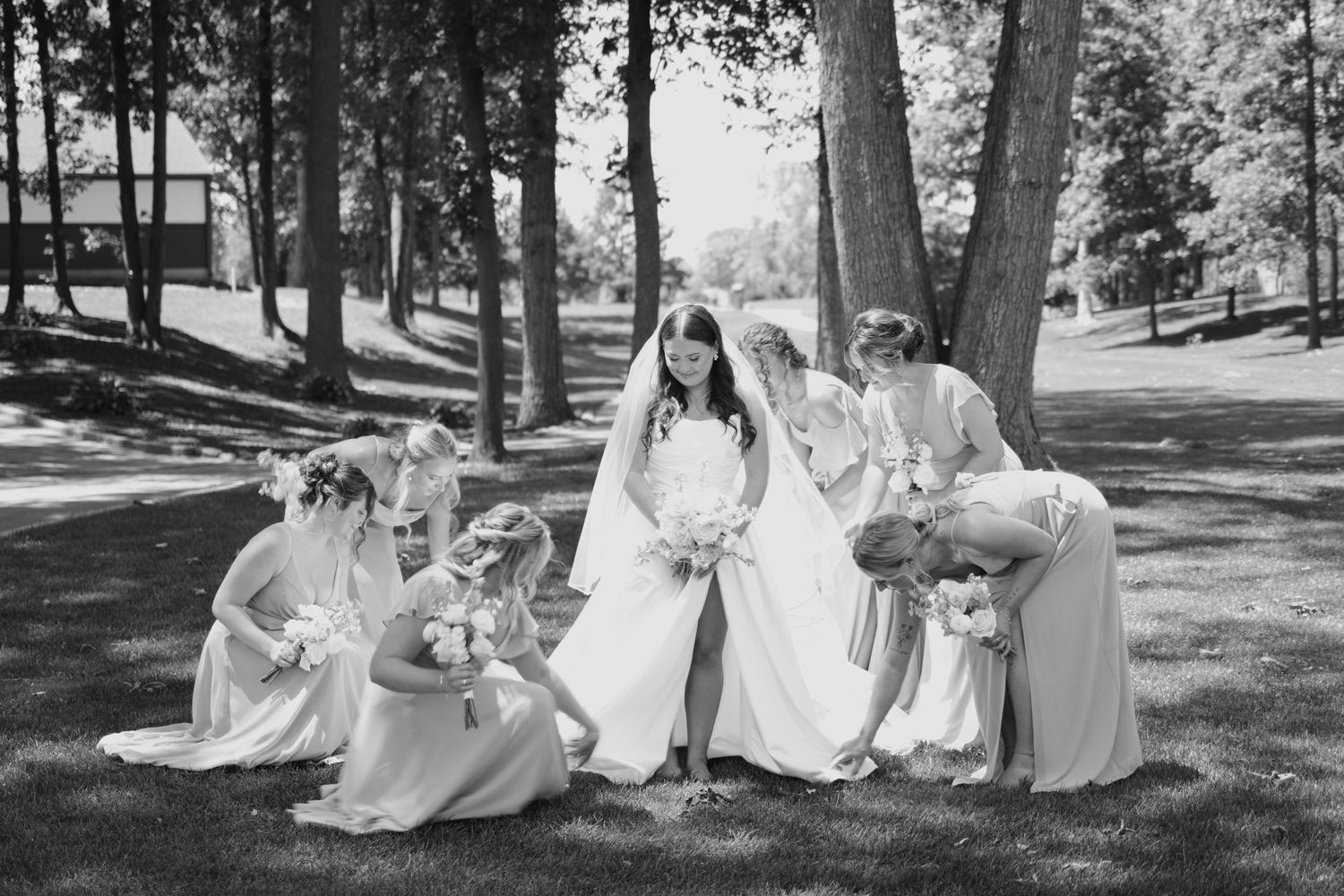 Black and white photo of a bride in a white gown and veil surrounded by bridesmaids in a sunny outdoor park setting.