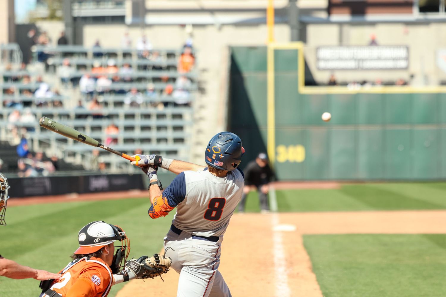 Baseball batter swings at pitch while catcher readies to receive at sunny ballpark field during daytime game.