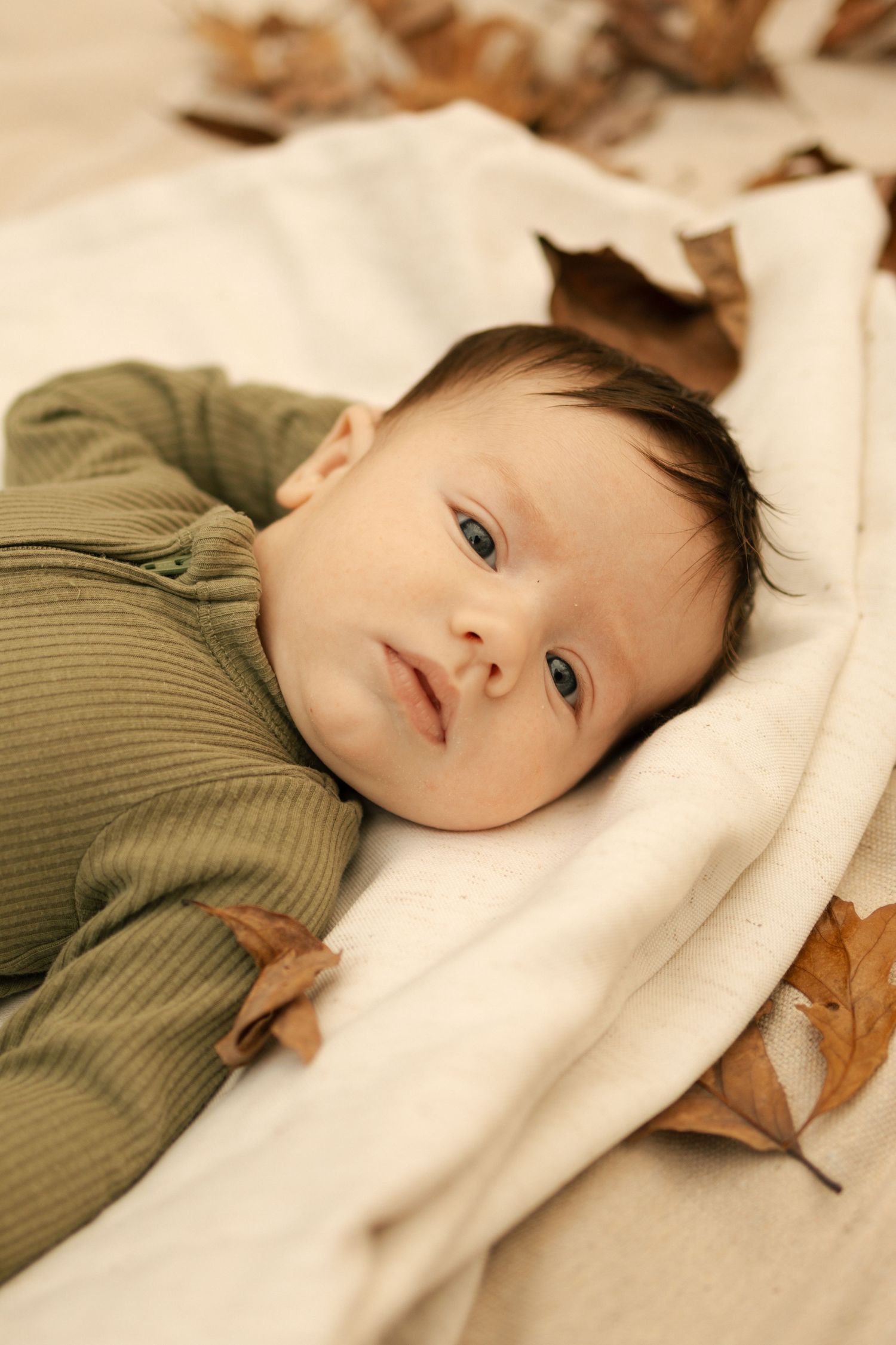 Newborn baby in green outfit lying on white blanket surrounded by autumn leaves, looking up with blue eyes.