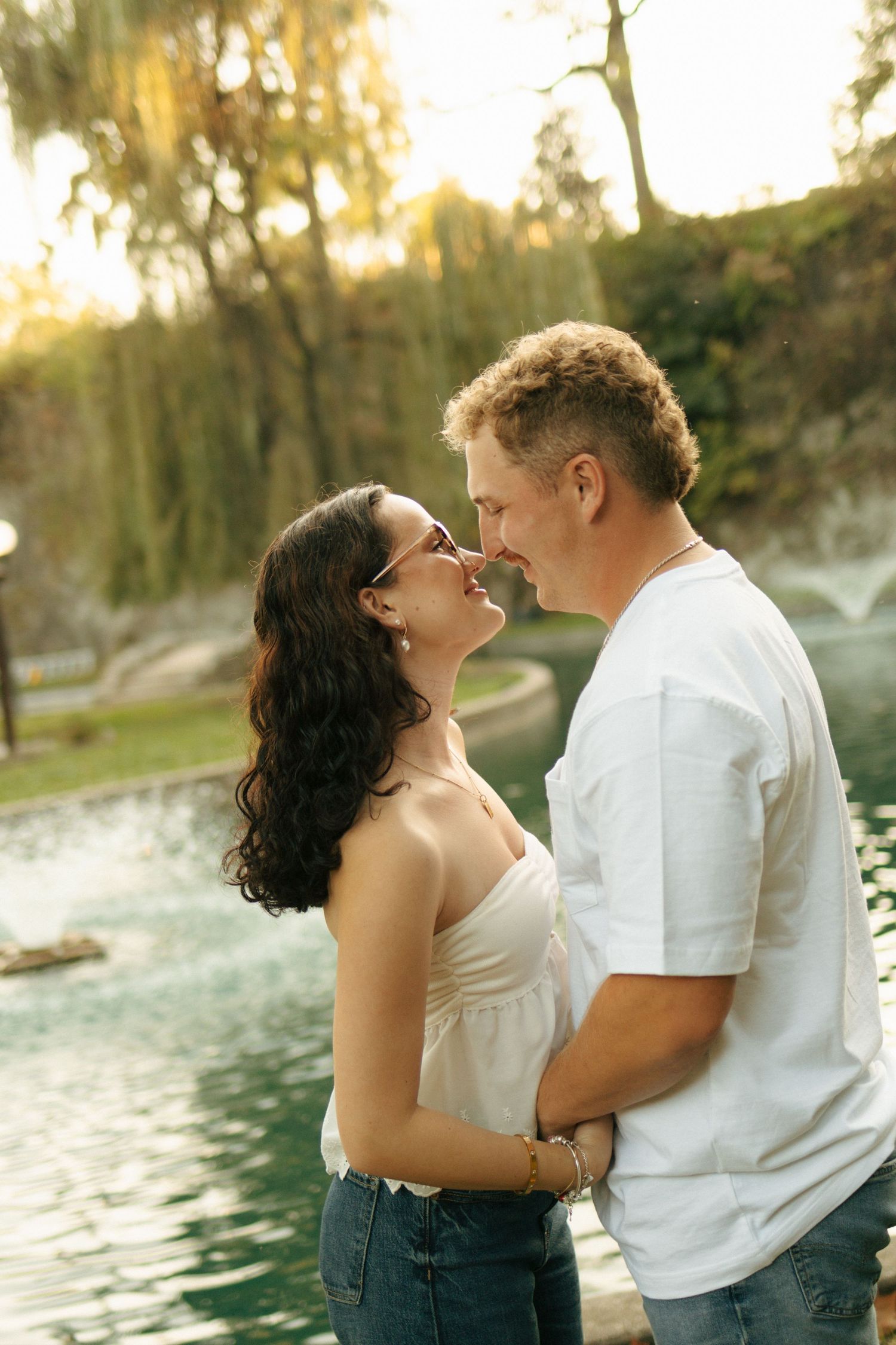 Romantic couple sharing a tender moment by a sunlit pond surrounded by lush green trees in a park.