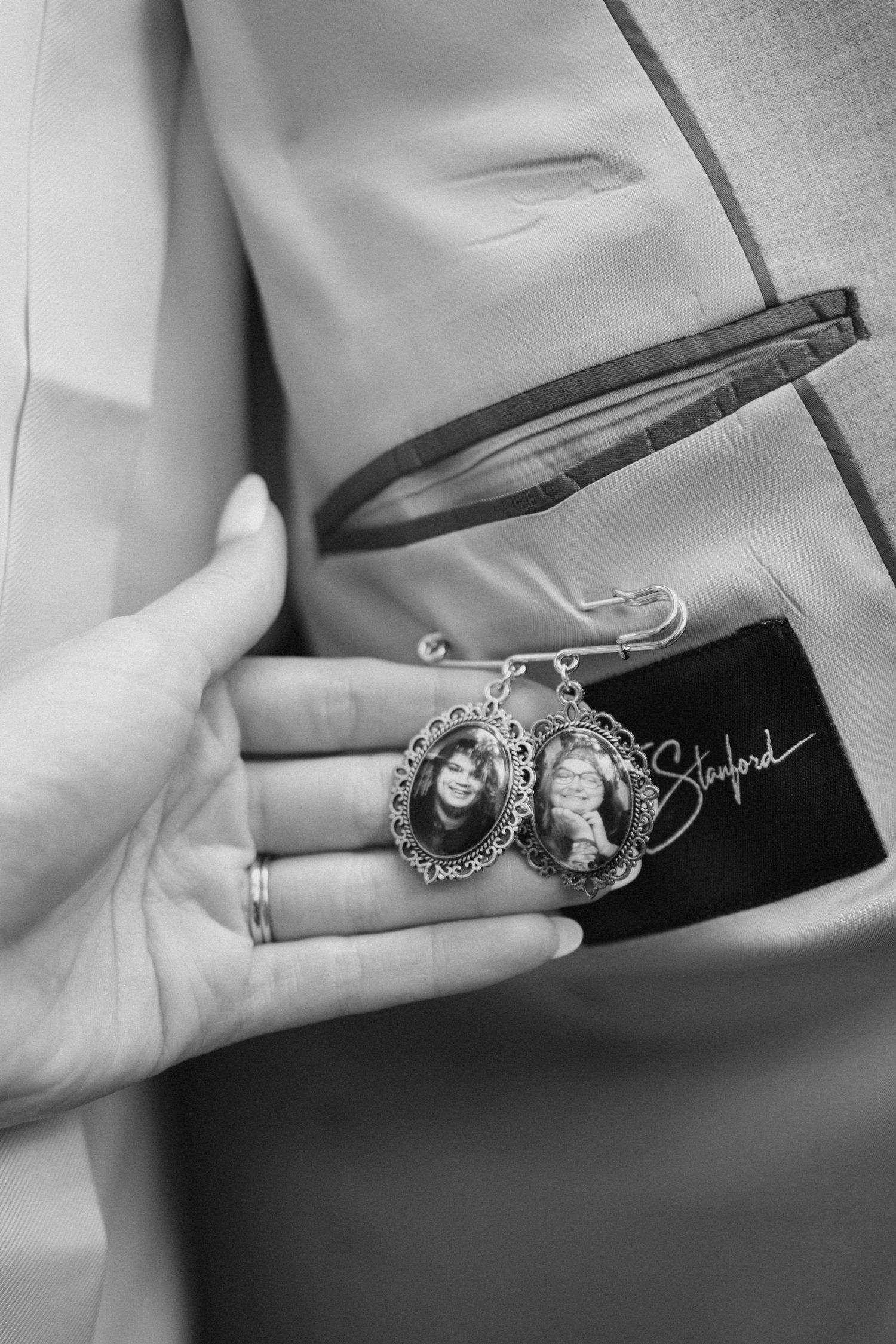 Black and white close-up of a hand holding two oval memorial photo charms pinned to a gray suit lapel.