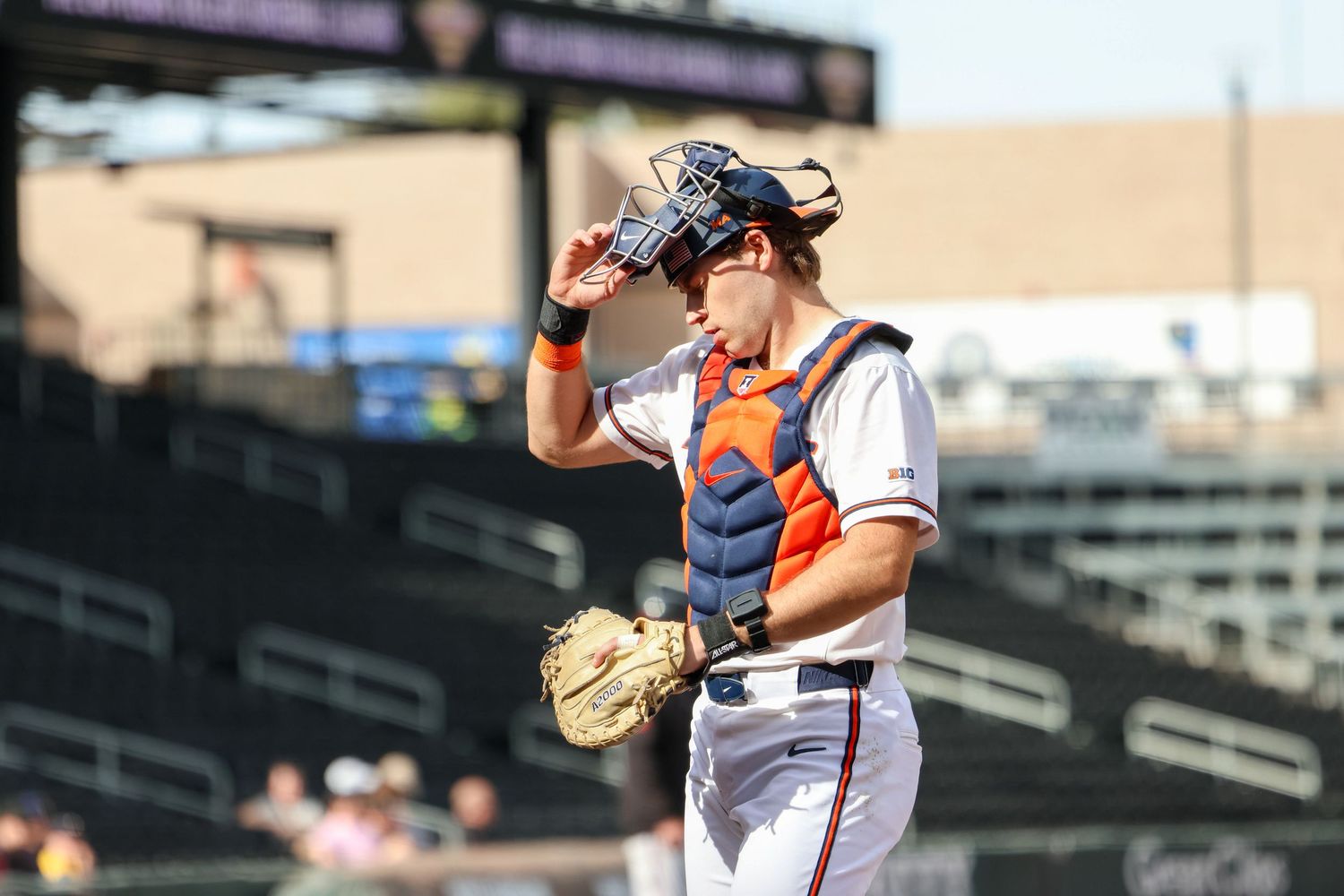 Baseball catcher in orange and white uniform wipes forehead during game at stadium.
