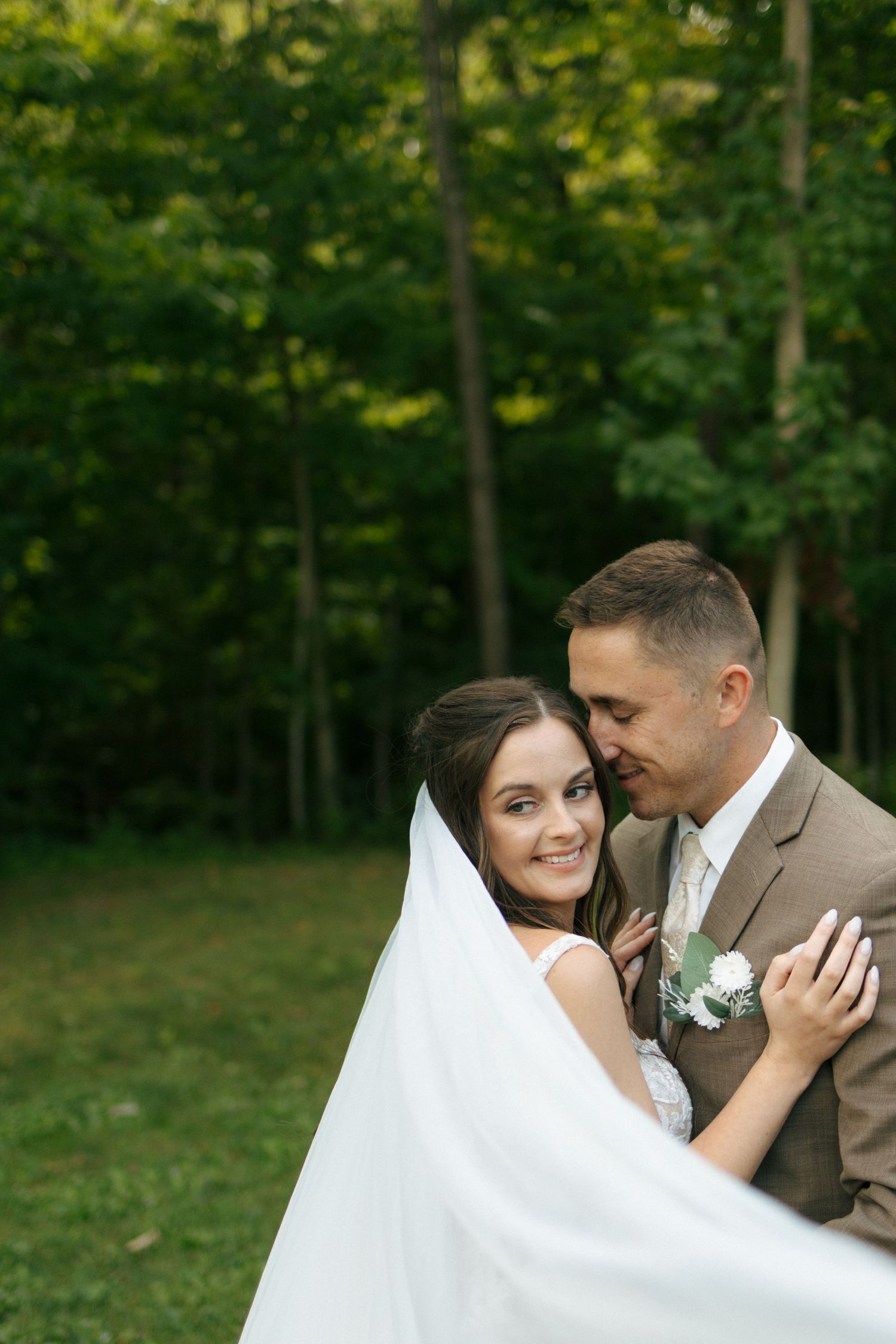 Romantic outdoor wedding photo with couple embracing under flowing white veil in lush green forest setting.