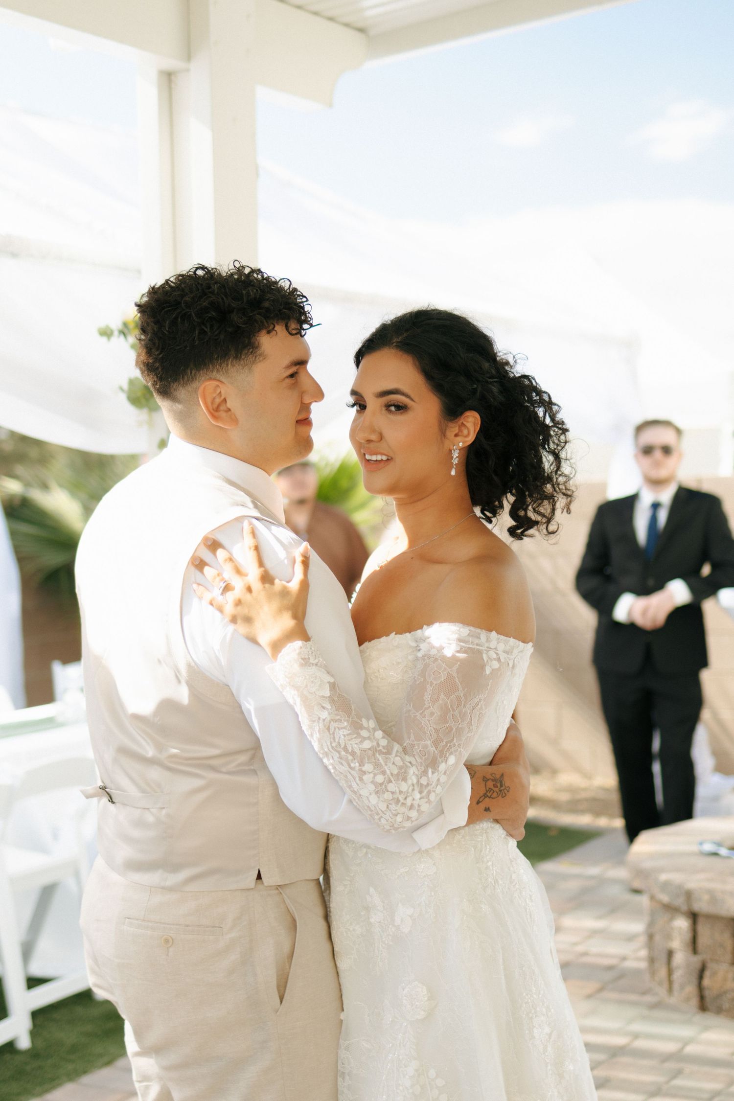 A couple shares an intimate moment during their wedding ceremony outdoors while an officiant stands in the background.