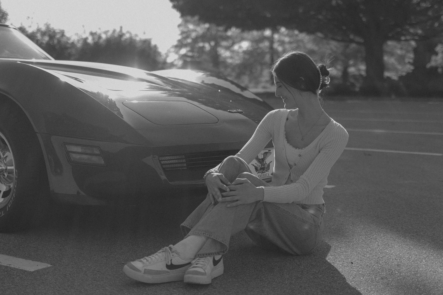Black and white photo of a young woman sitting on pavement beside a classic sports car in a parking lot.