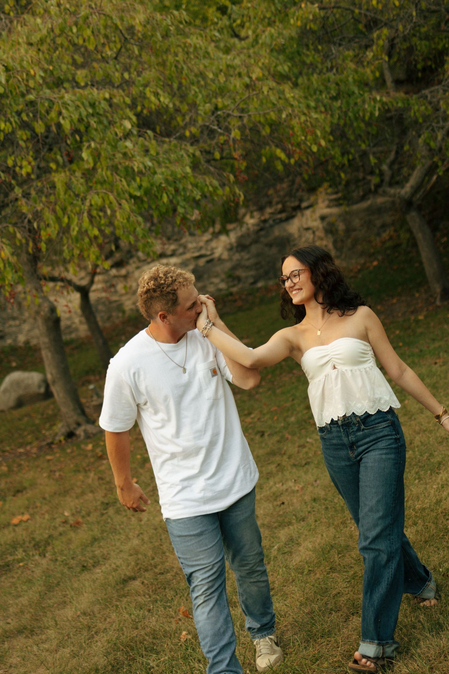 A couple walks hand-in-hand through a lush green park, smiling at each other in casual white tops and jeans.