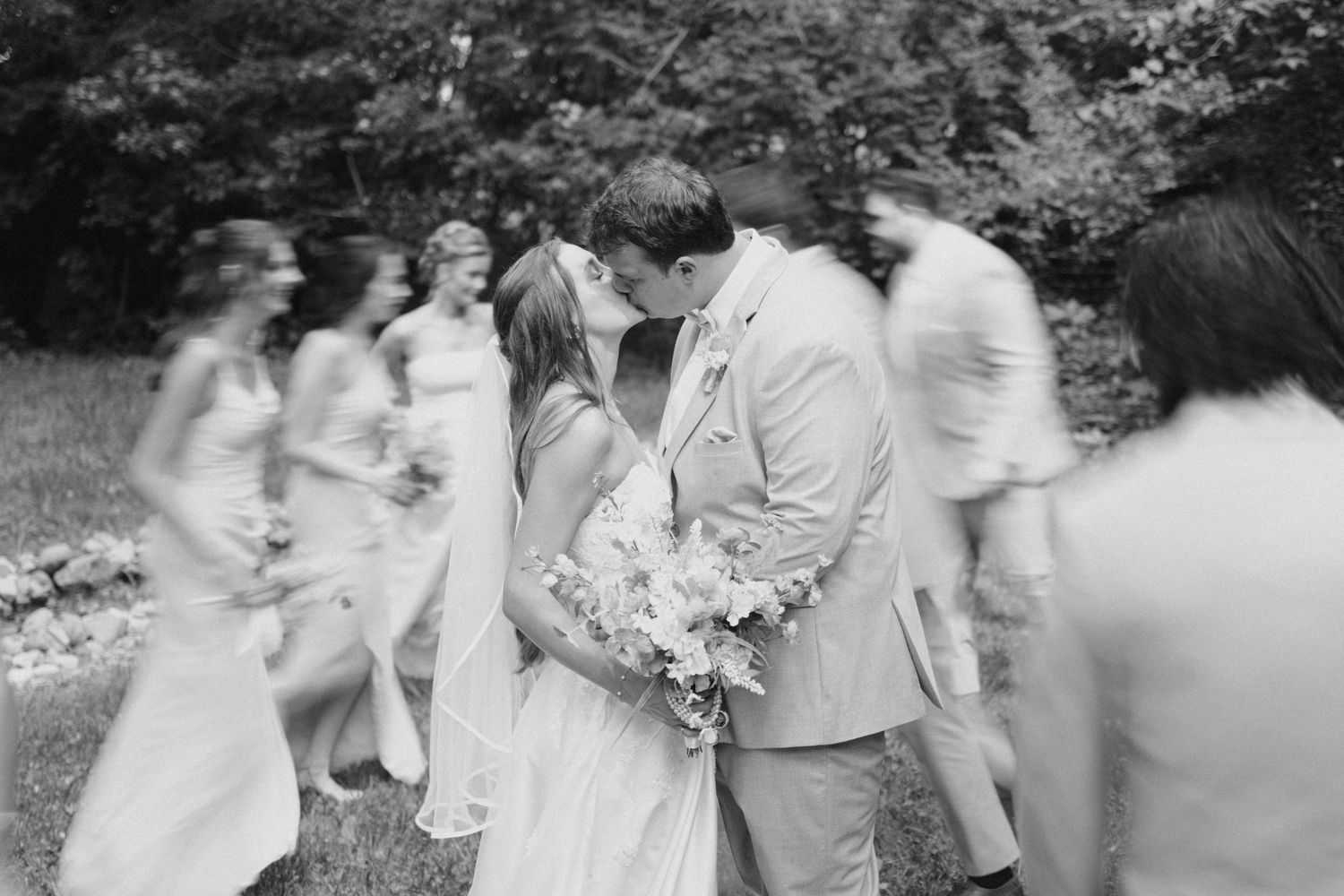 Black and white photo of bride and groom kissing while wedding party walks past in blur.