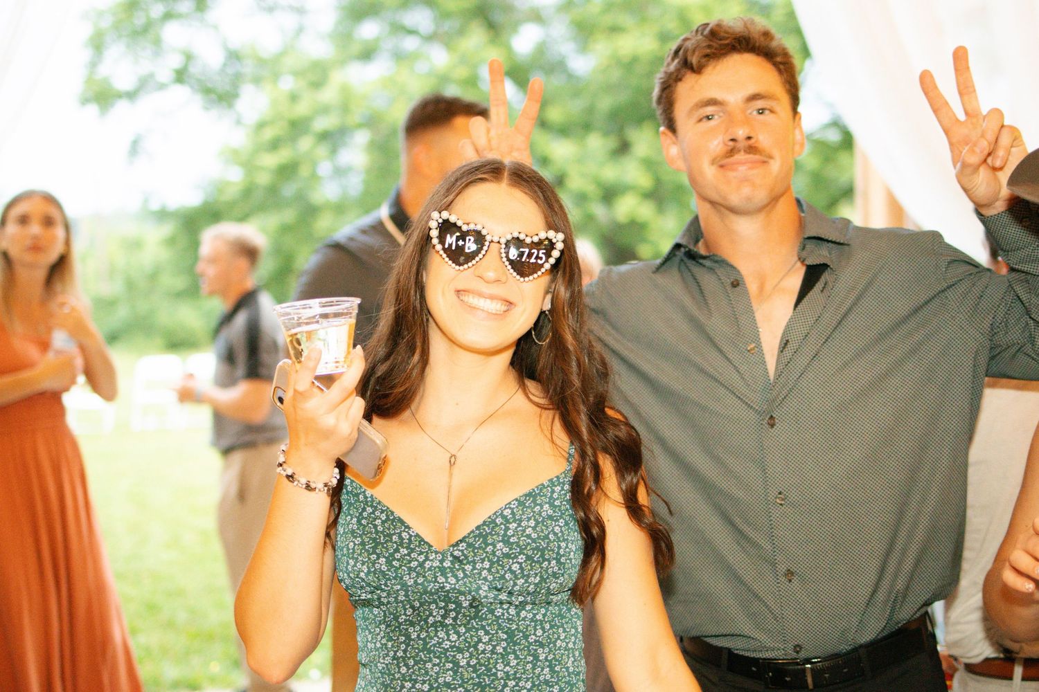 Wedding guest in green dress and heart sunglasses poses with peace sign man at outdoor reception.