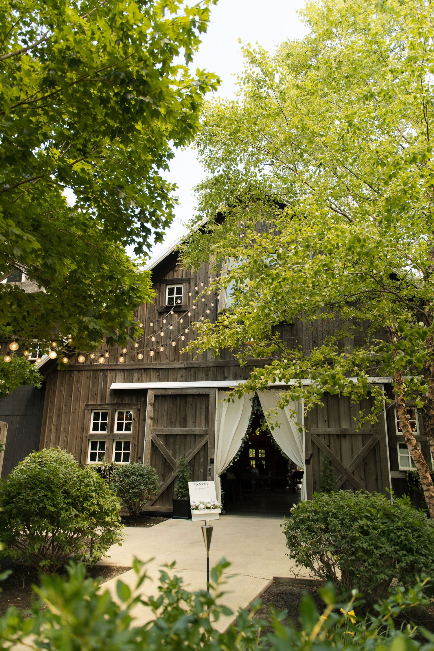Rustic wooden barn venue with string lights, white curtain doorway, lush green trees, and manicured shrubs for an outdoor event.