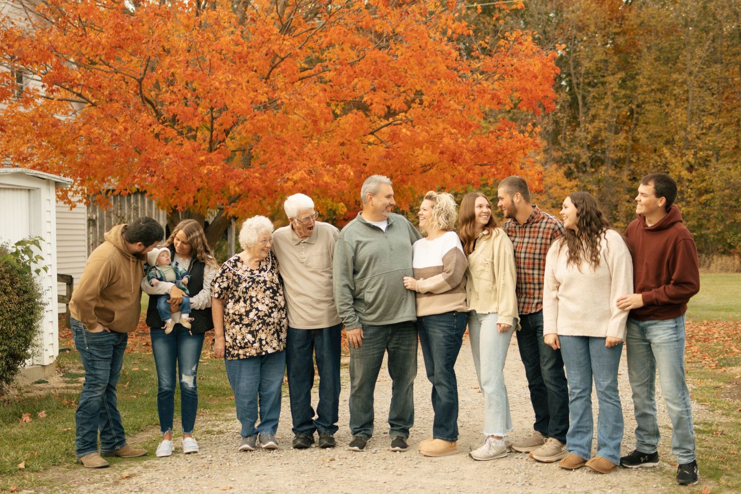 Multi-generational family gathered outdoors in autumn, smiling together near vibrant orange fall foliage trees.