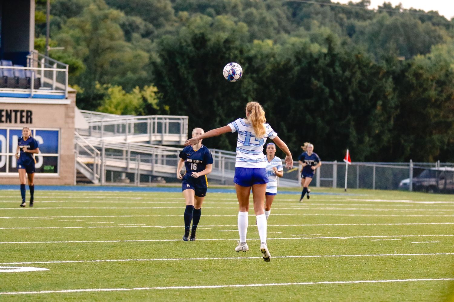 Two soccer players leap to compete for the ball during a match on an outdoor field with bleachers visible in background.