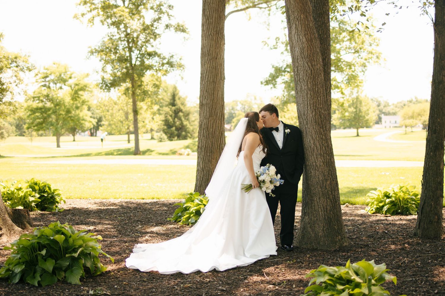Bride and groom share a romantic kiss leaning against a tree in a lush green outdoor wedding setting.