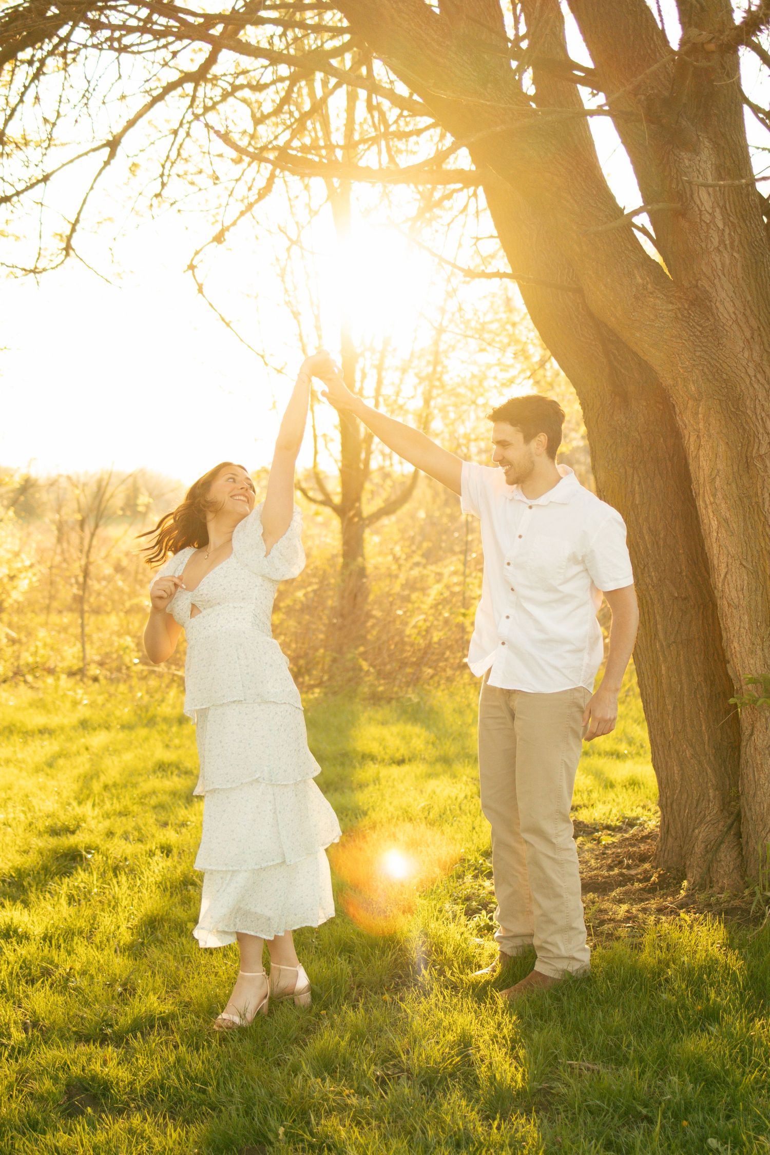 Couple dancing outdoors in golden sunset light near a tree on green grass, woman in white dress, man in white shirt.
