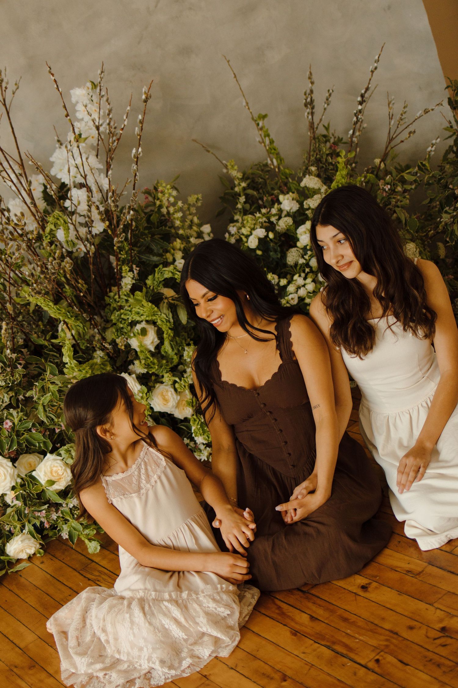 Three women in elegant dresses sit together surrounded by lush white floral arrangements on a wooden floor.