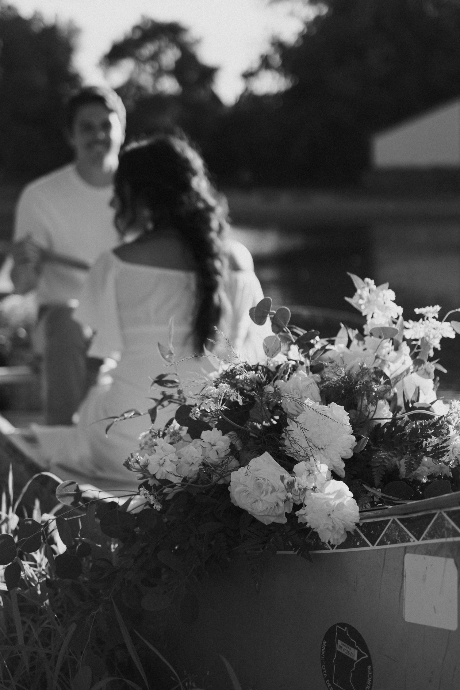 Black and white photo of a floral arrangement in foreground with a couple sharing a romantic moment in the background.
