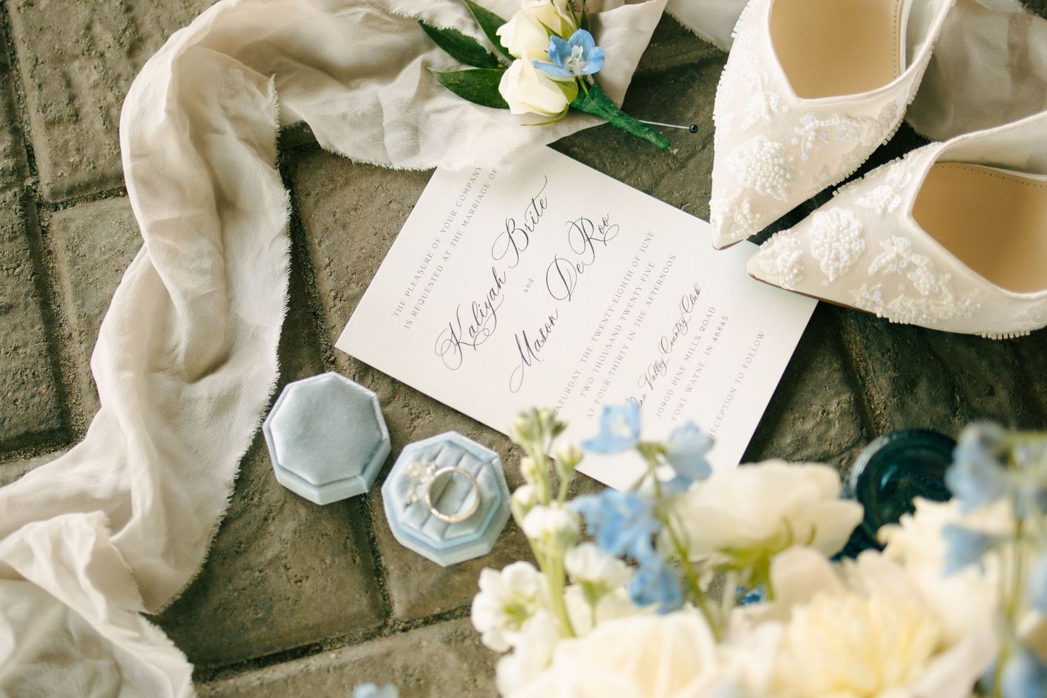 Elegant wedding flat lay with lace bridal heels, floral bouquet, blue ring box, and calligraphy invitation on stone surface.
