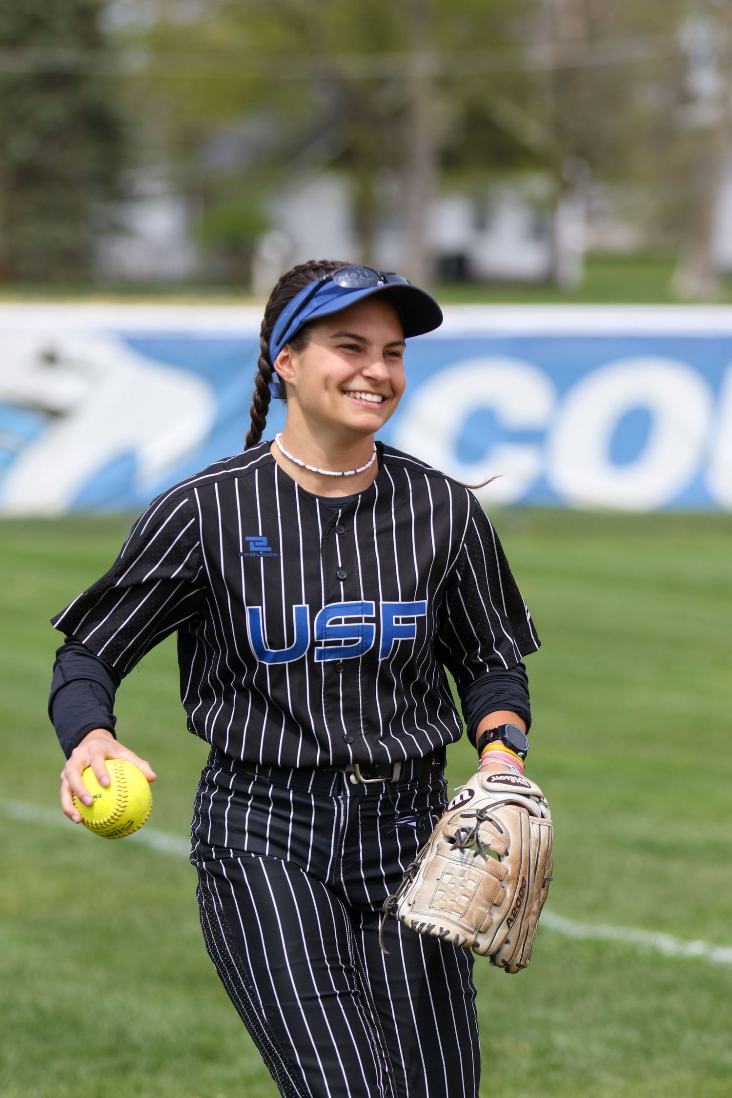 Softball player wearing pinstriped USA uniform smiles while holding yellow ball and glove on grassy field during practice.