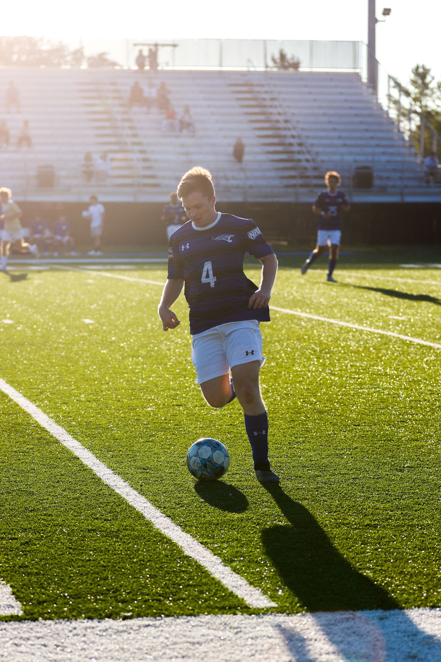 Soccer player dribbles ball on green field during sunset with stadium bleachers in background.