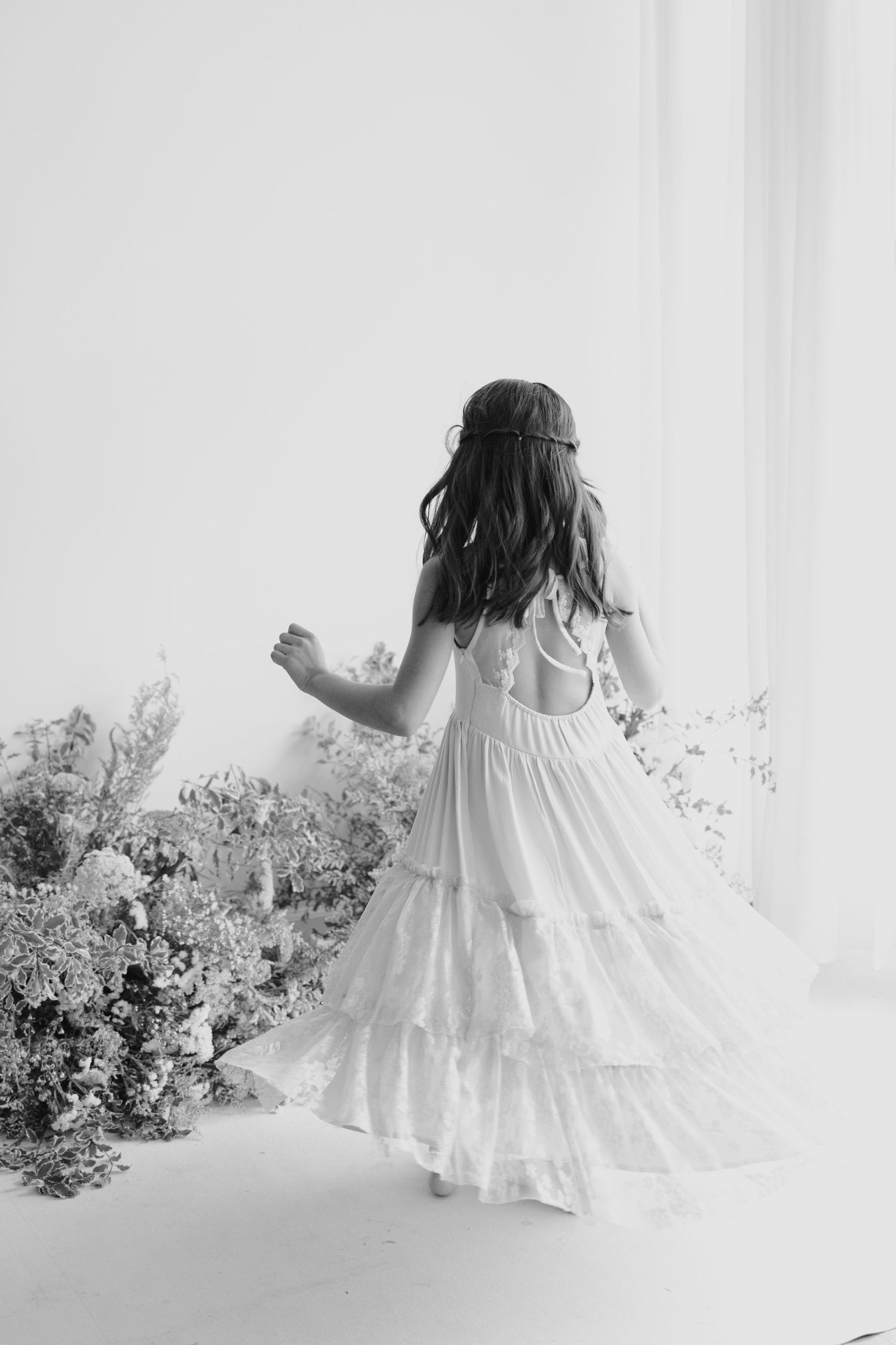 Black and white photo of a girl in a tiered white lace dress dancing near flowers in a bright studio.