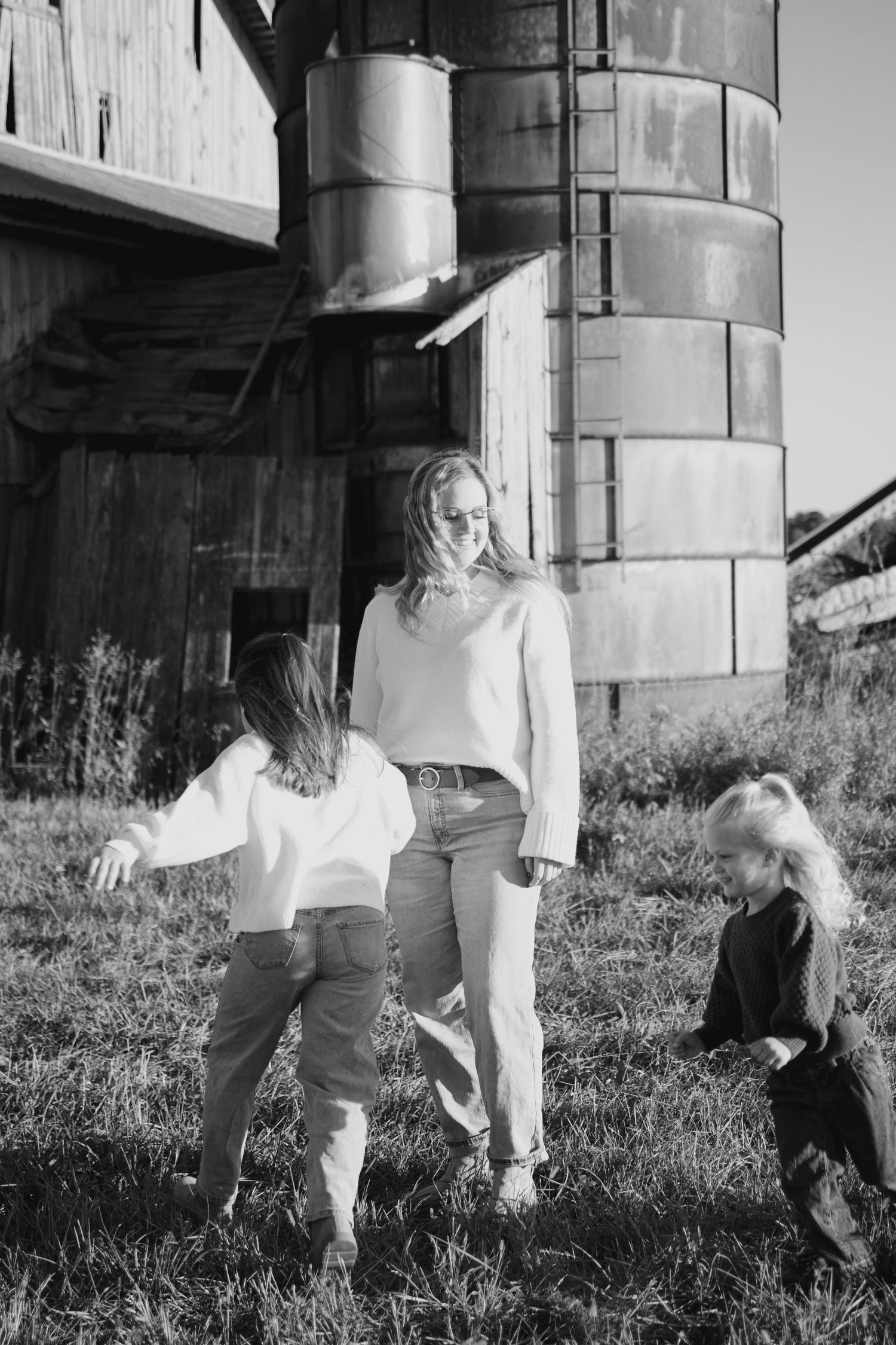 Black and white photo of a mother and two daughters playing outdoors near a rustic barn and grain silo on a farm.