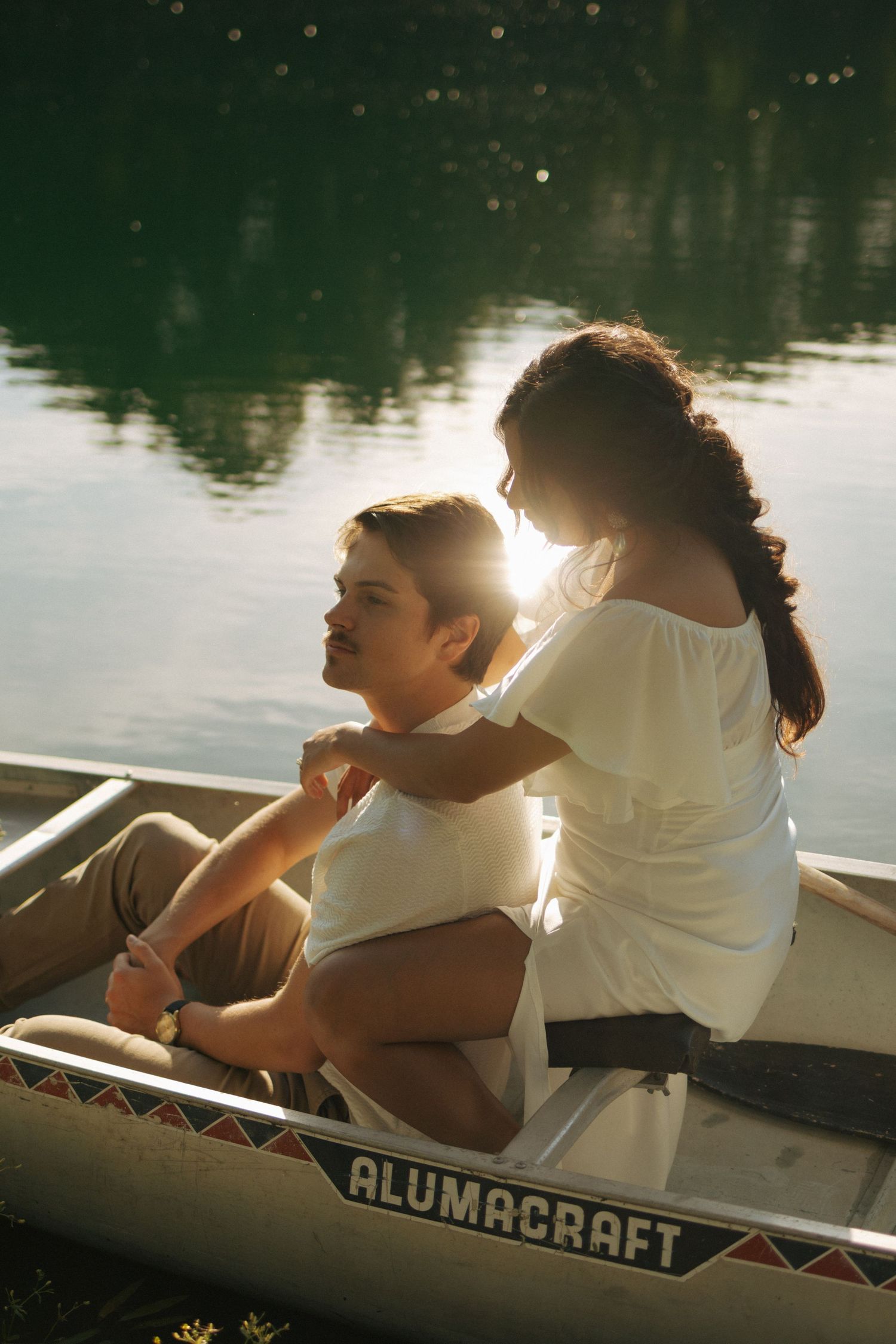 Couple sitting in an Alumacraft canoe on a sunlit lake, woman in white dress touching mans shoulders romantically.