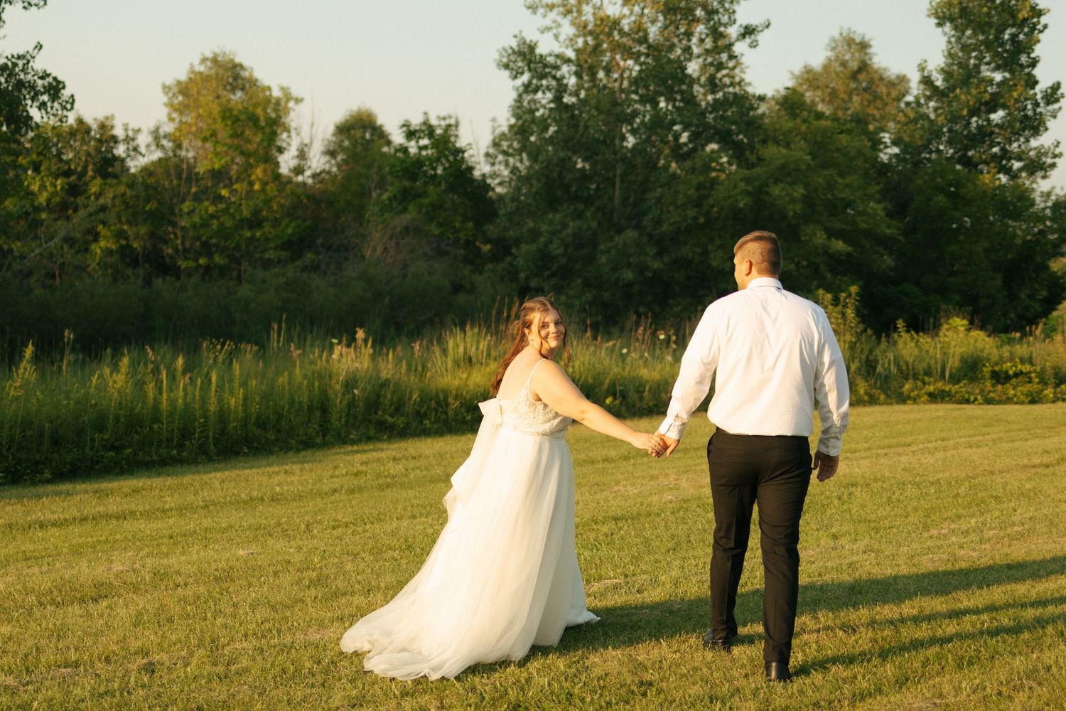 A bride and groom walk hand in hand across a sunlit green meadow surrounded by lush trees during golden hour.