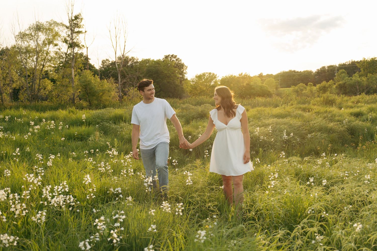Couple holding hands walking through a sunny green meadow with wildflowers at golden hour.