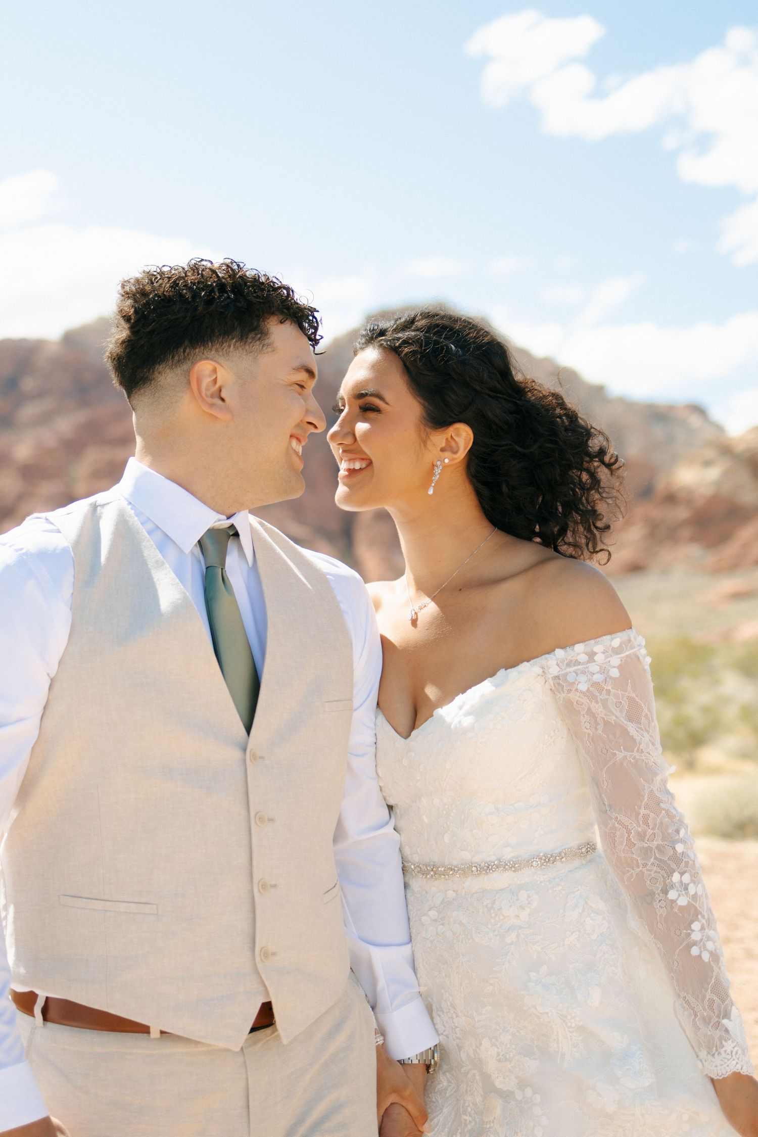 A couple shares a joyful intimate moment in off-white wedding attire against a desert landscape backdrop.