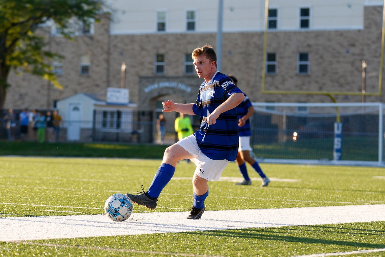 Soccer player in navy uniform dribbles ball across sunny grass field with stone building in background.